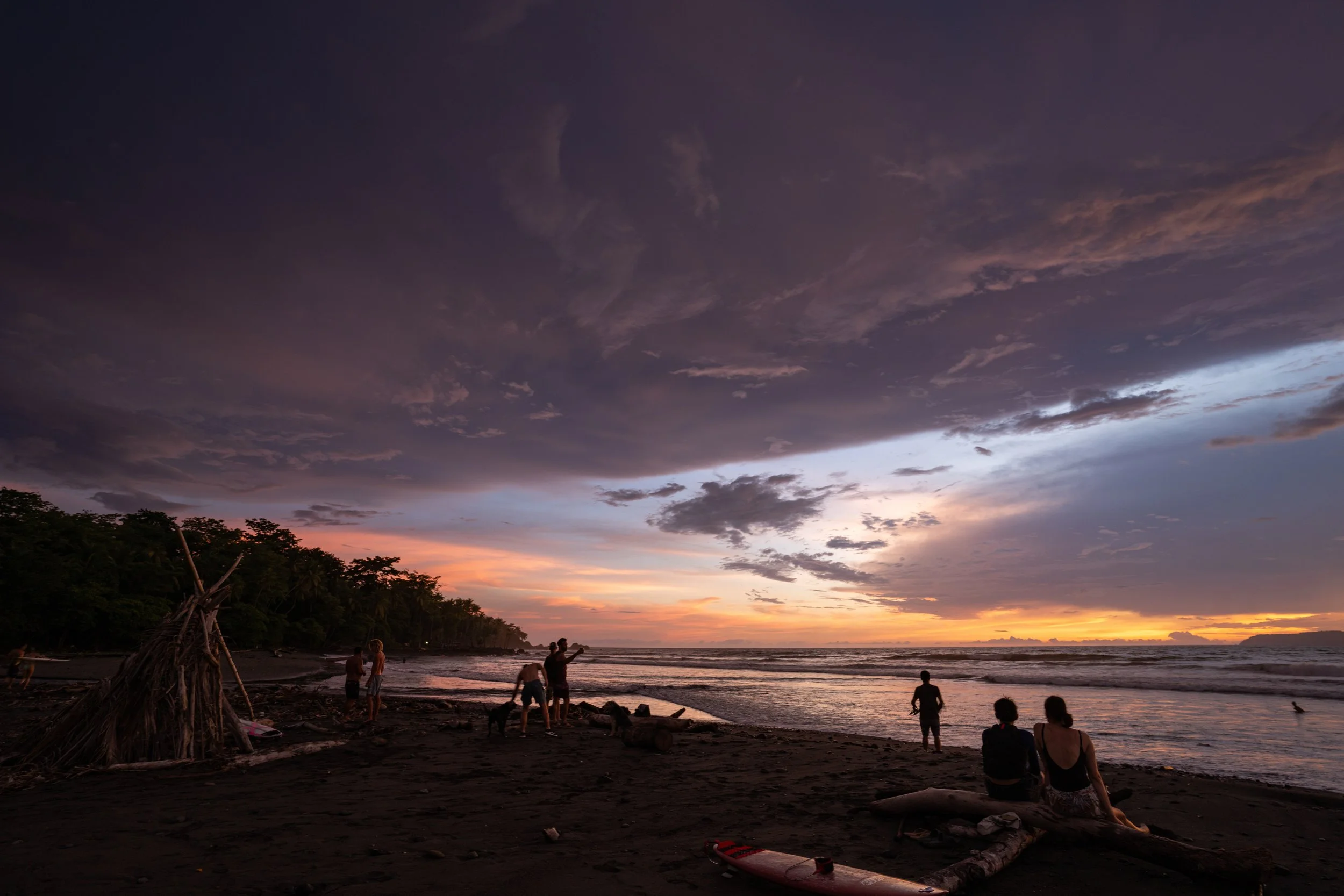 A beach scene during sunset with a dark sky overhead. People are gathered on the beach, some standing and talking, others sitting, with a surfboard and driftwood on the sand. Trees are visible in the background along the shoreline.