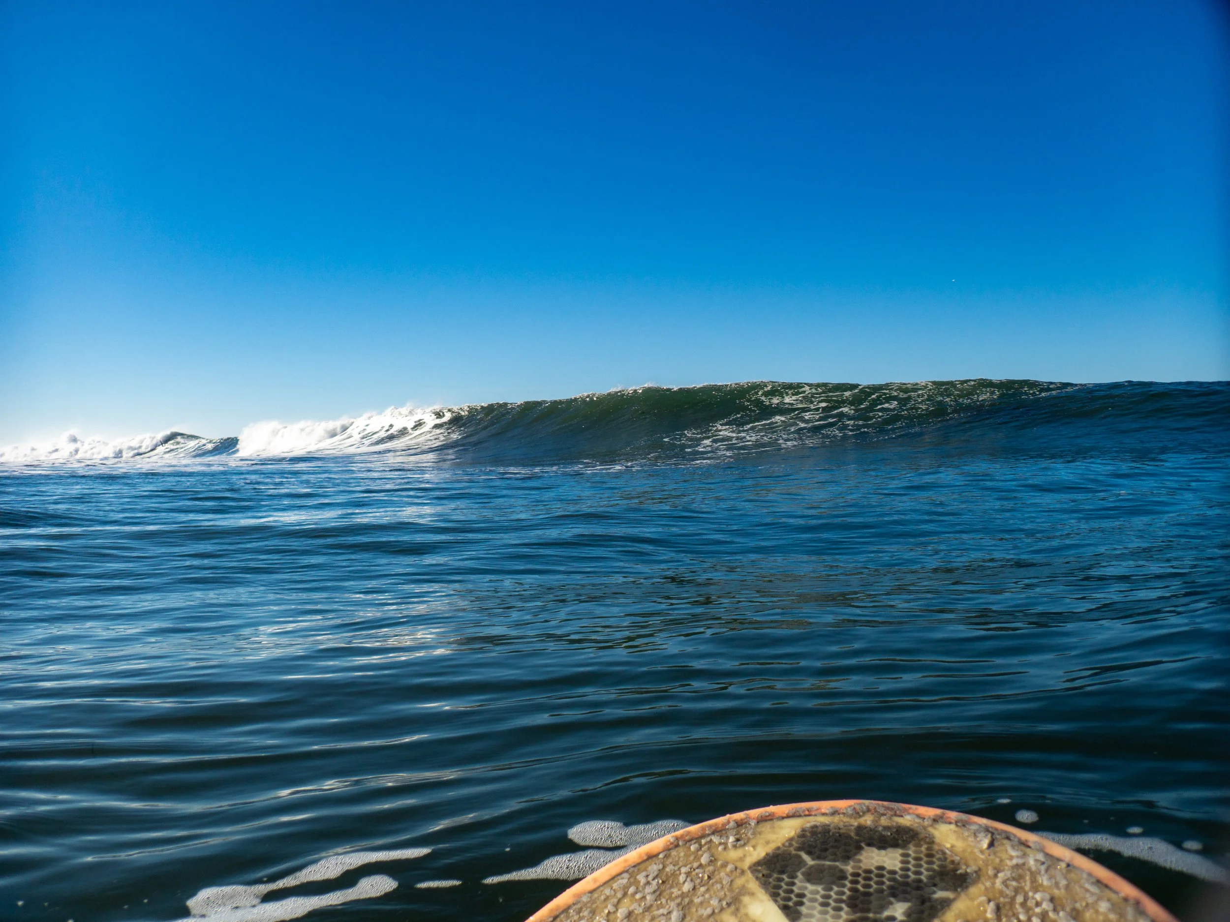 View of the ocean with a large wave in the distance and part of a surfboard in the foreground on the water.