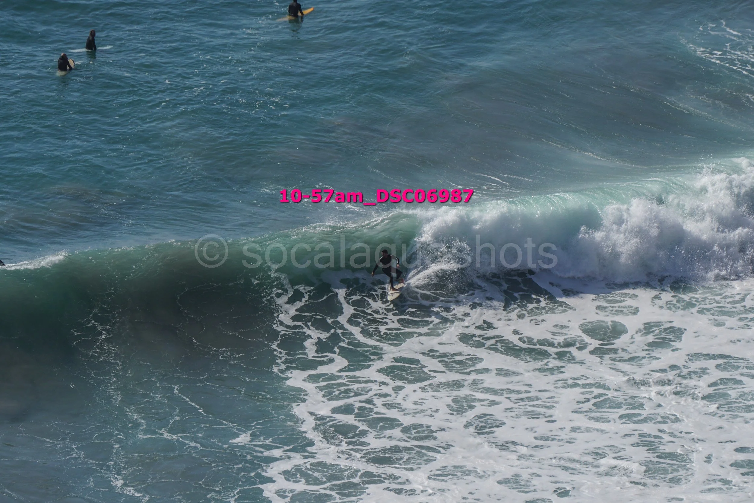 Surfer riding a wave in the ocean, with several people swimming or surfboarding in the background.