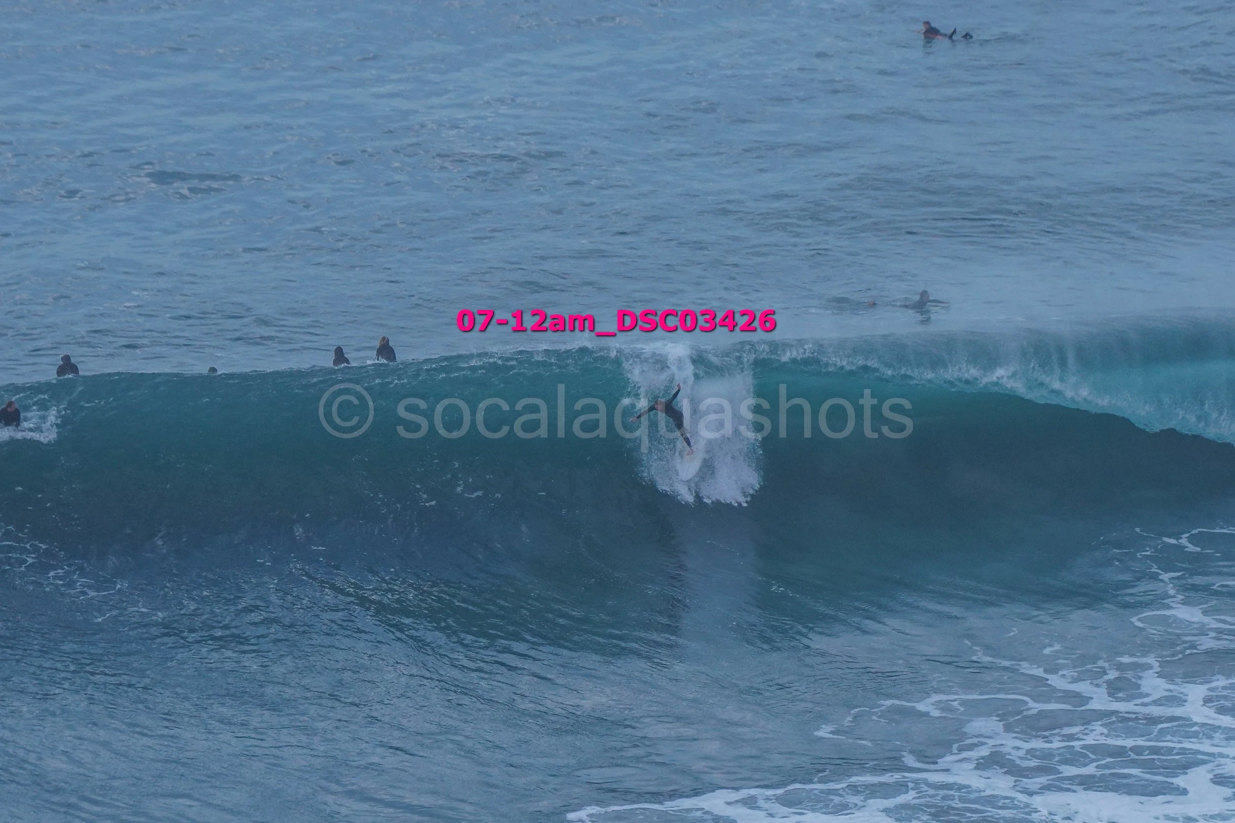 A group of surfers in the ocean, some riding a large wave, others waiting in the water, with one surfer mid-ride on a wave.
