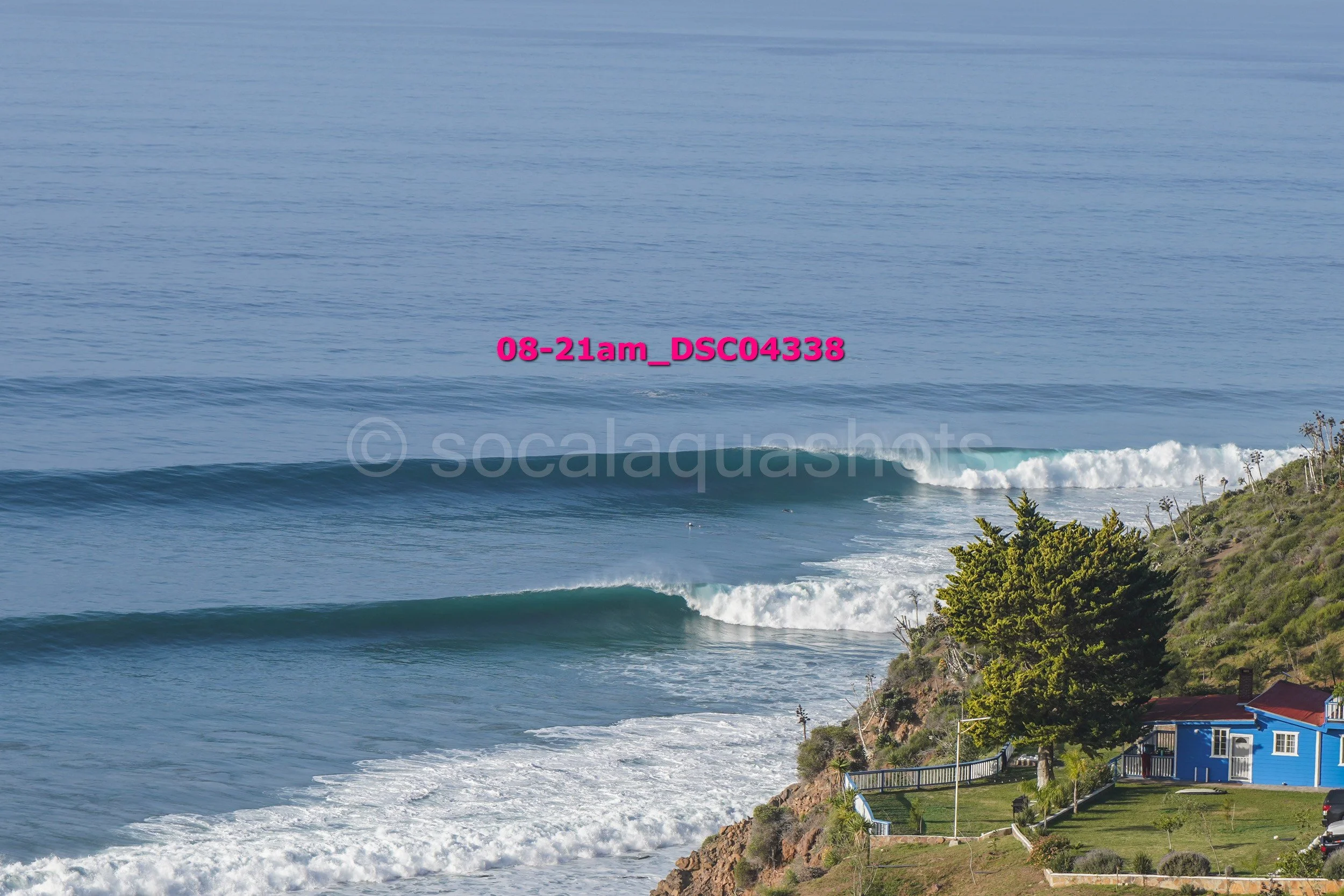 Coastal scene with large ocean waves, green hill with trees, blue house, and parked cars.