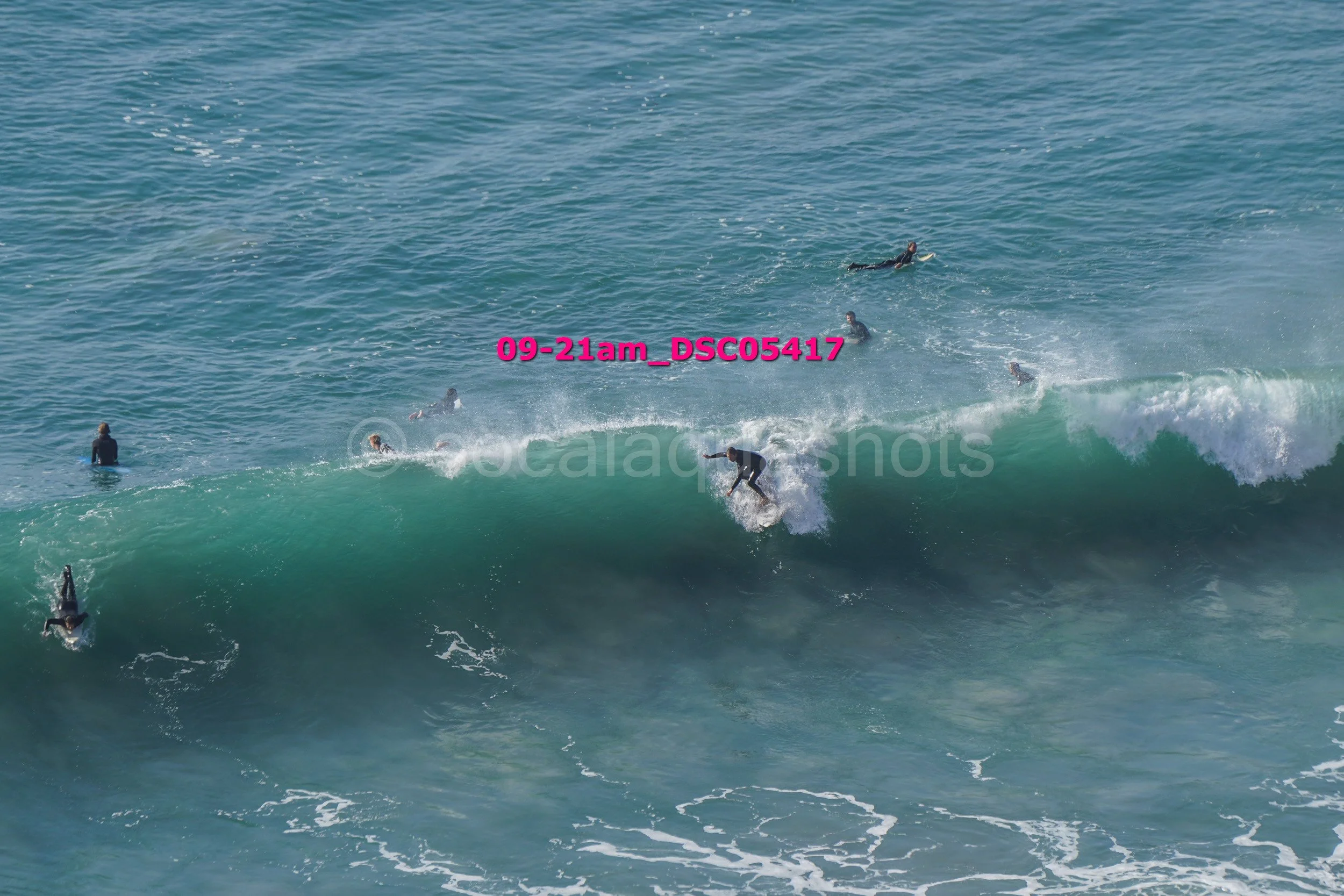Surfers riding a large wave in the ocean, with some surfers waiting and others paddling out in the background.