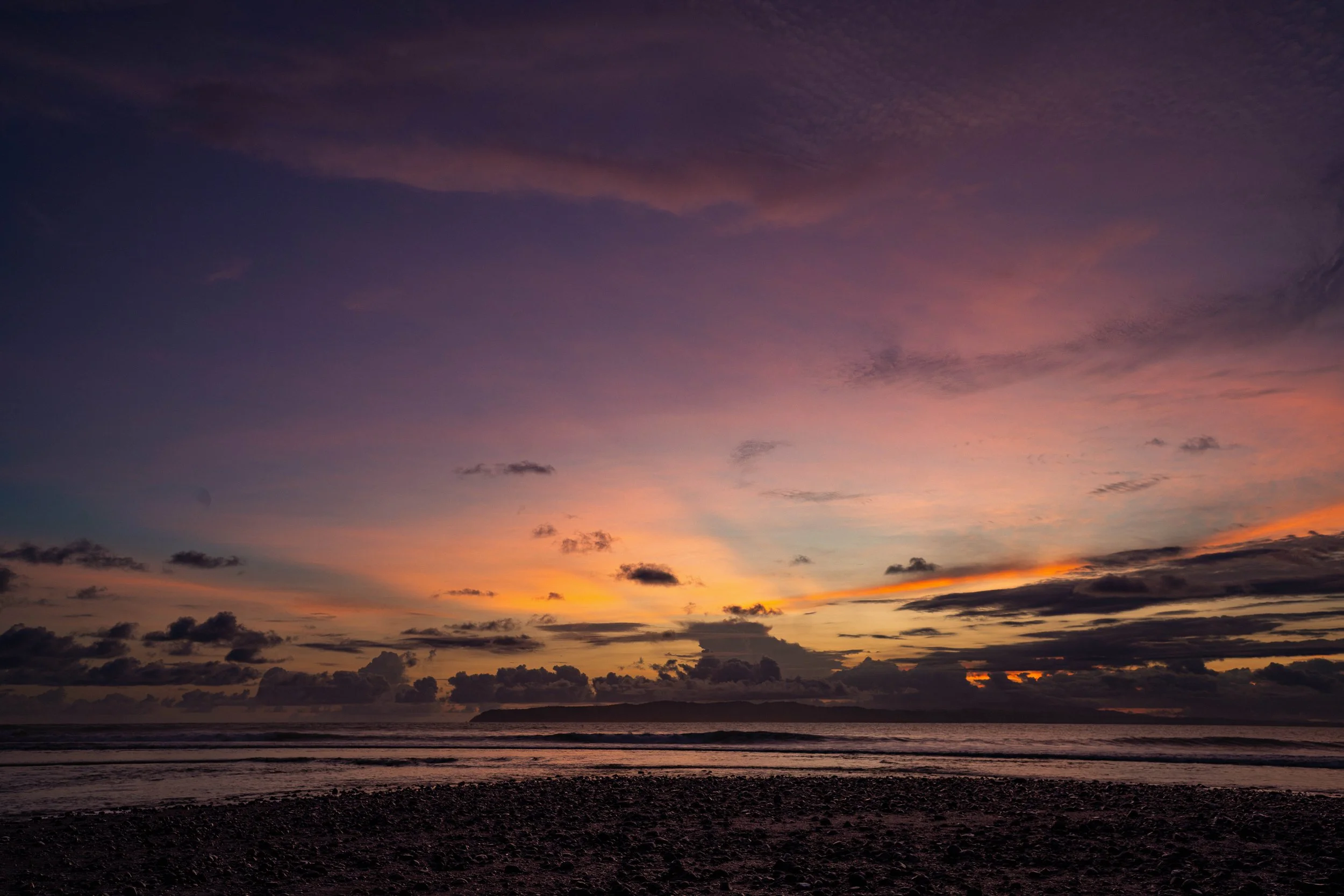 A colorful sunset over the ocean with a dark silhouetted shoreline and scattered clouds in the sky.