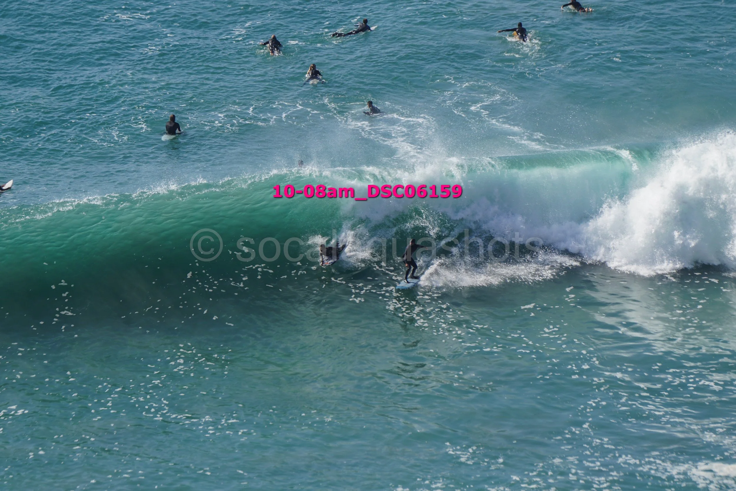 Group of surfers in the ocean, some riding a large wave while others paddle or wait in the water.