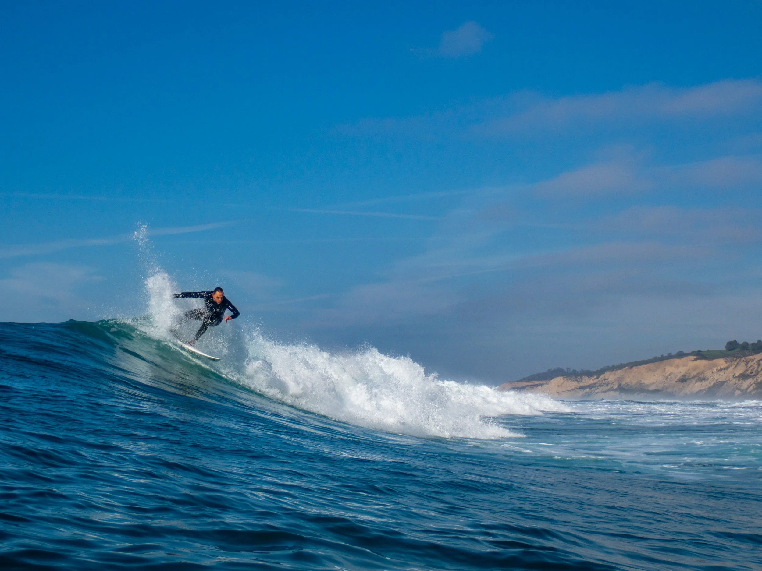 A person surfing on a wave in the ocean with a coastal landscape in the background.