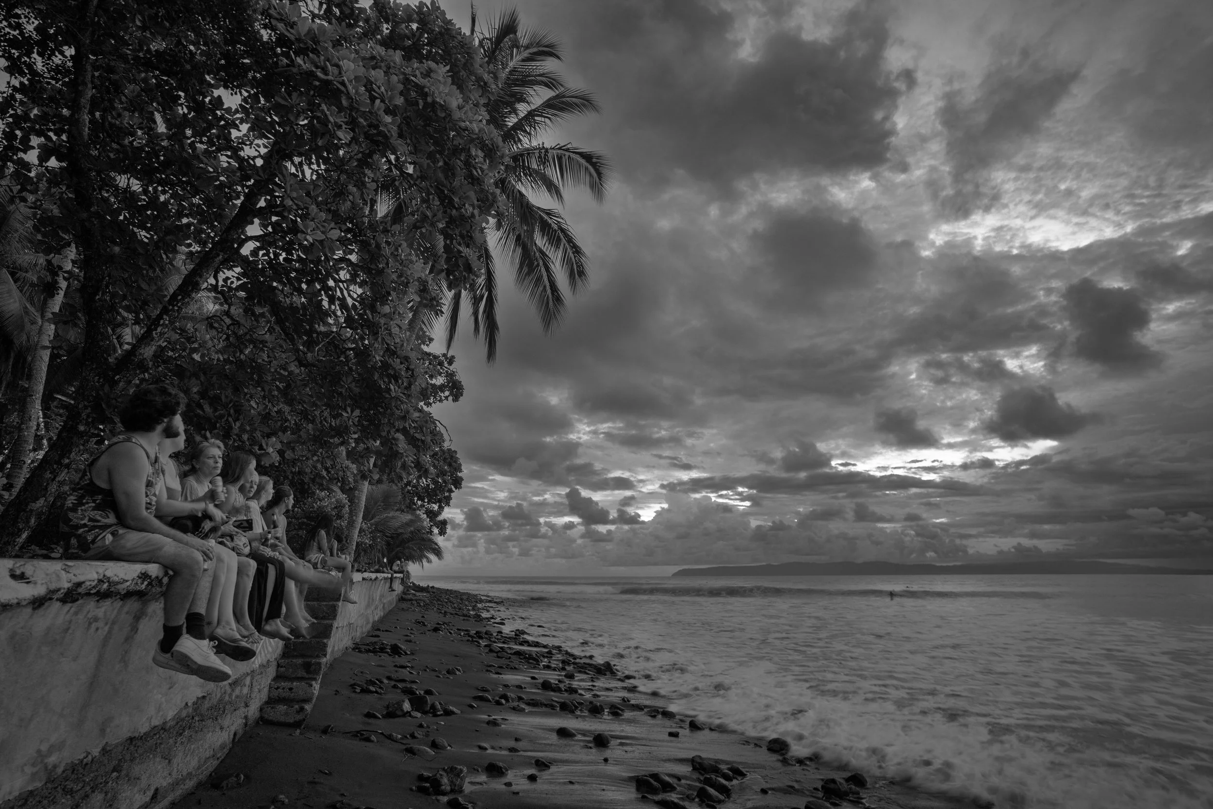 People sitting on a stone wall by the beach, with palm trees and cloudy sky in black and white.
