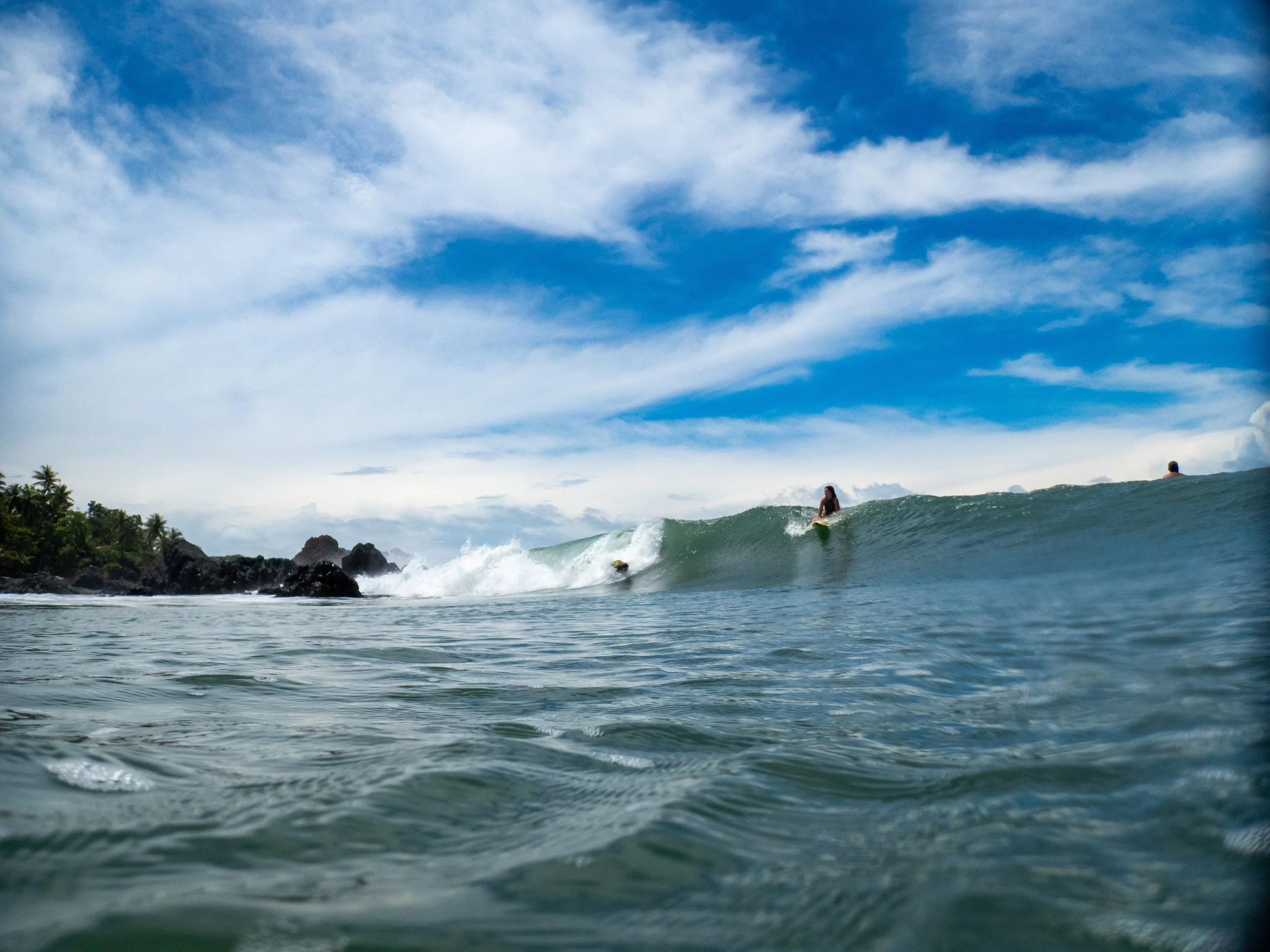 People surfing in the ocean with a shoreline, rocks, and trees on the left under a blue sky with clouds.
