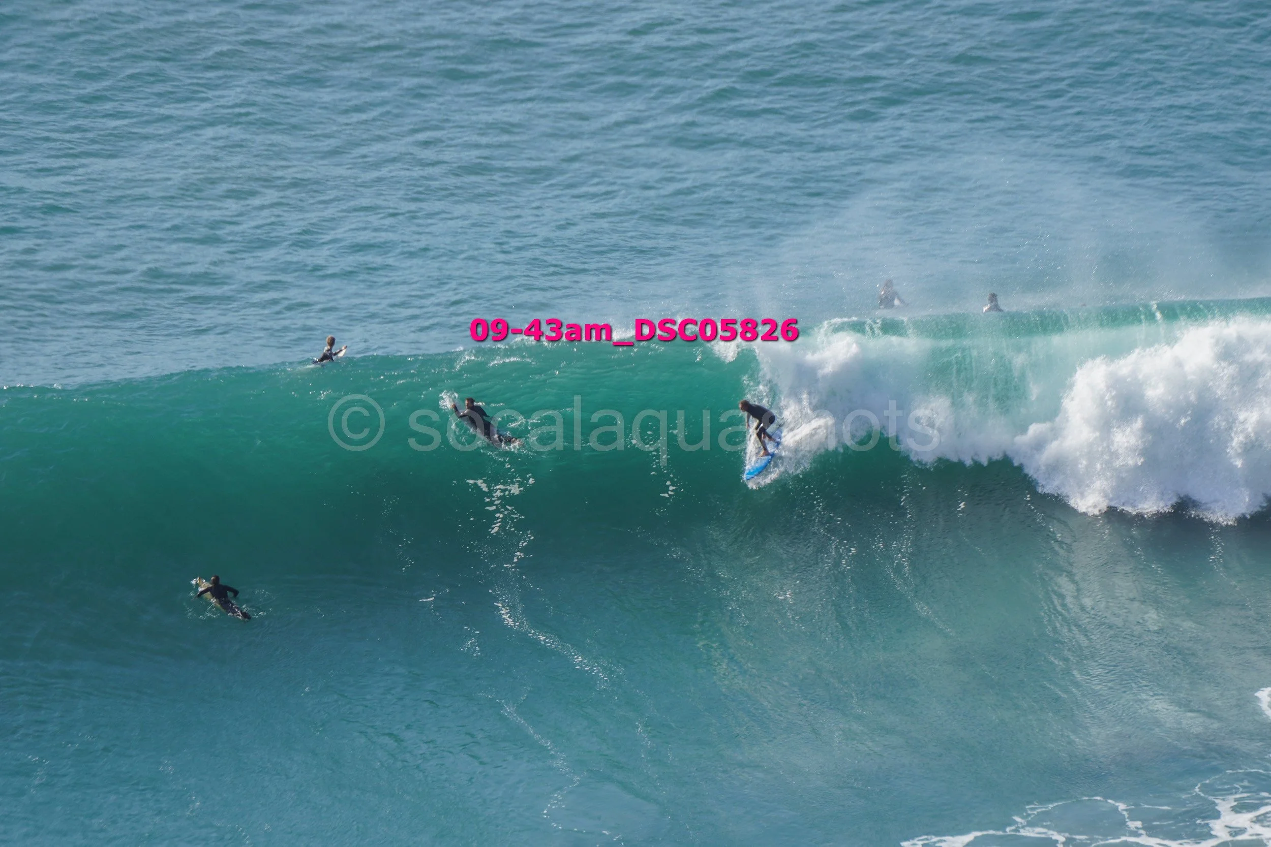 Surfers riding a large ocean wave with others in the water, some waiting and others paddling, under a clear sky.