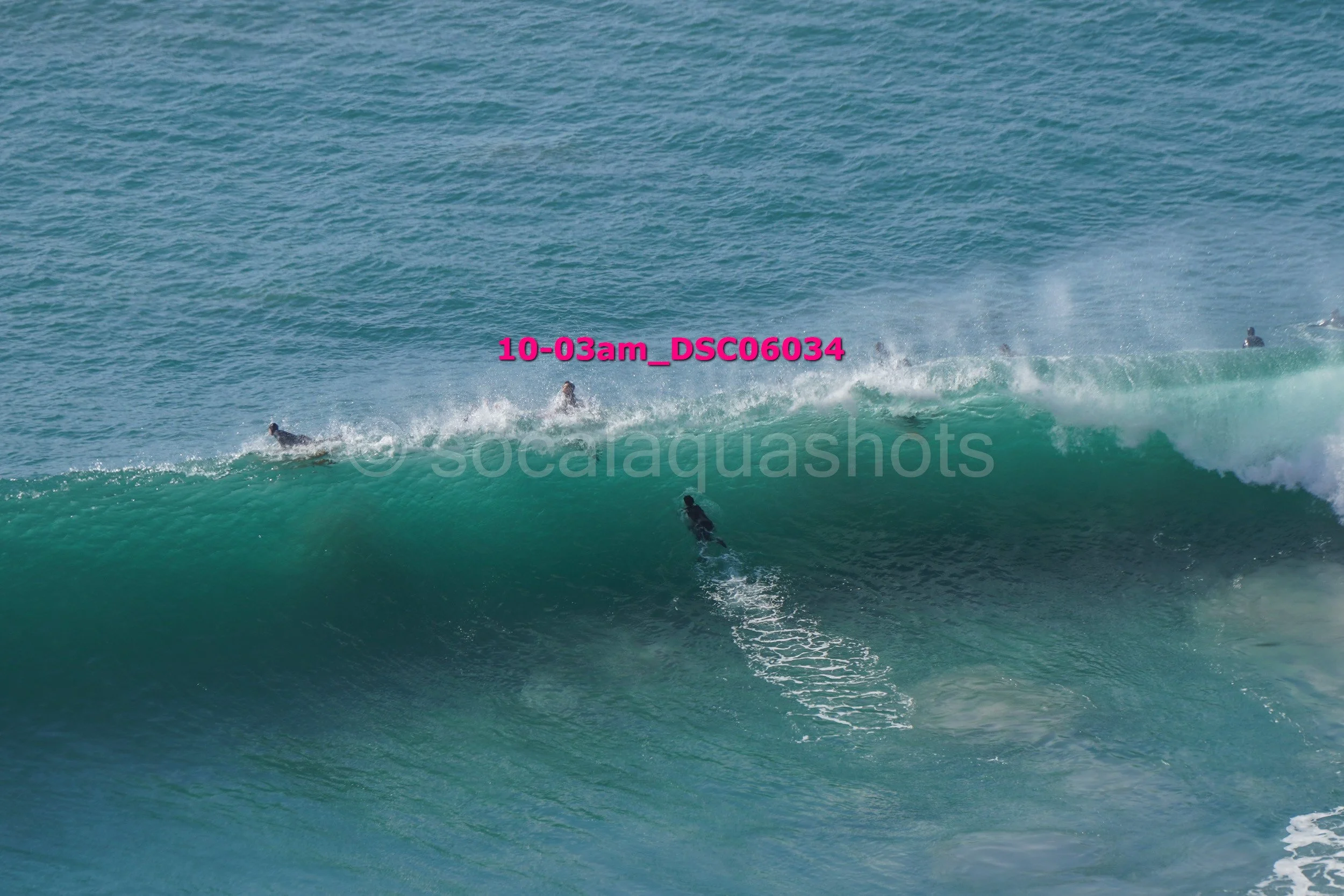 Surfers ride a large rolling wave in the ocean, some are on their boards, others are falling or preparing to surf.