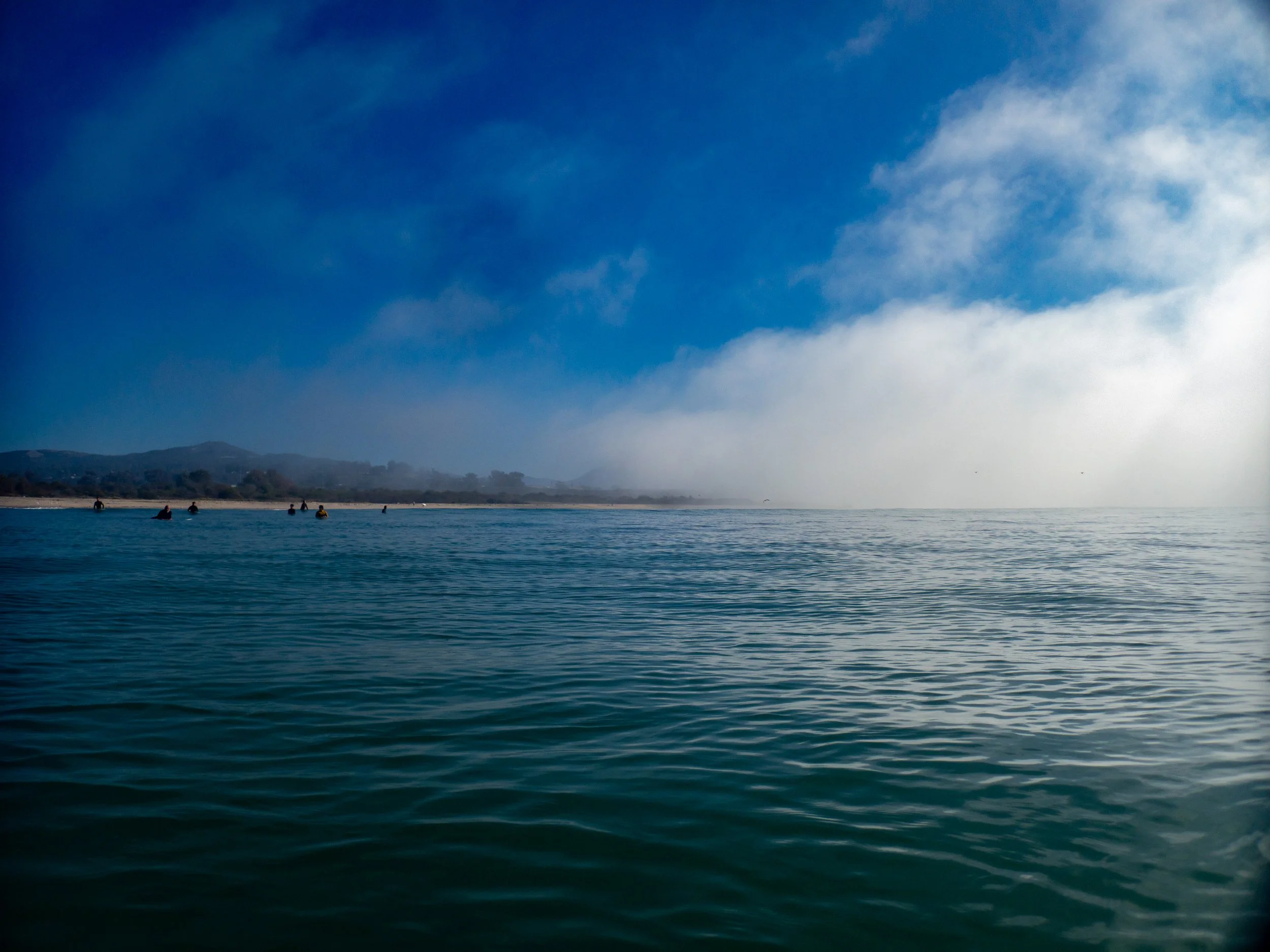 A serene ocean scene with a calm water surface, distant shoreline, and a partly cloudy sky with a patch of fog or mist over the horizon.