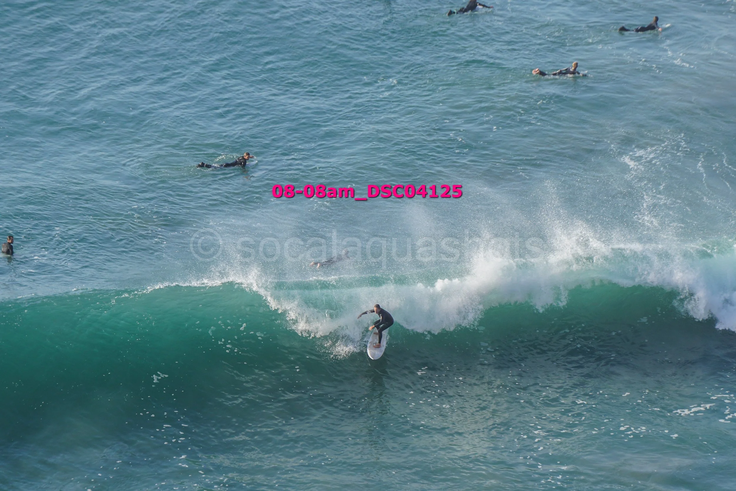 Surfer riding a wave with multiple people swimming and surfing in the background.
