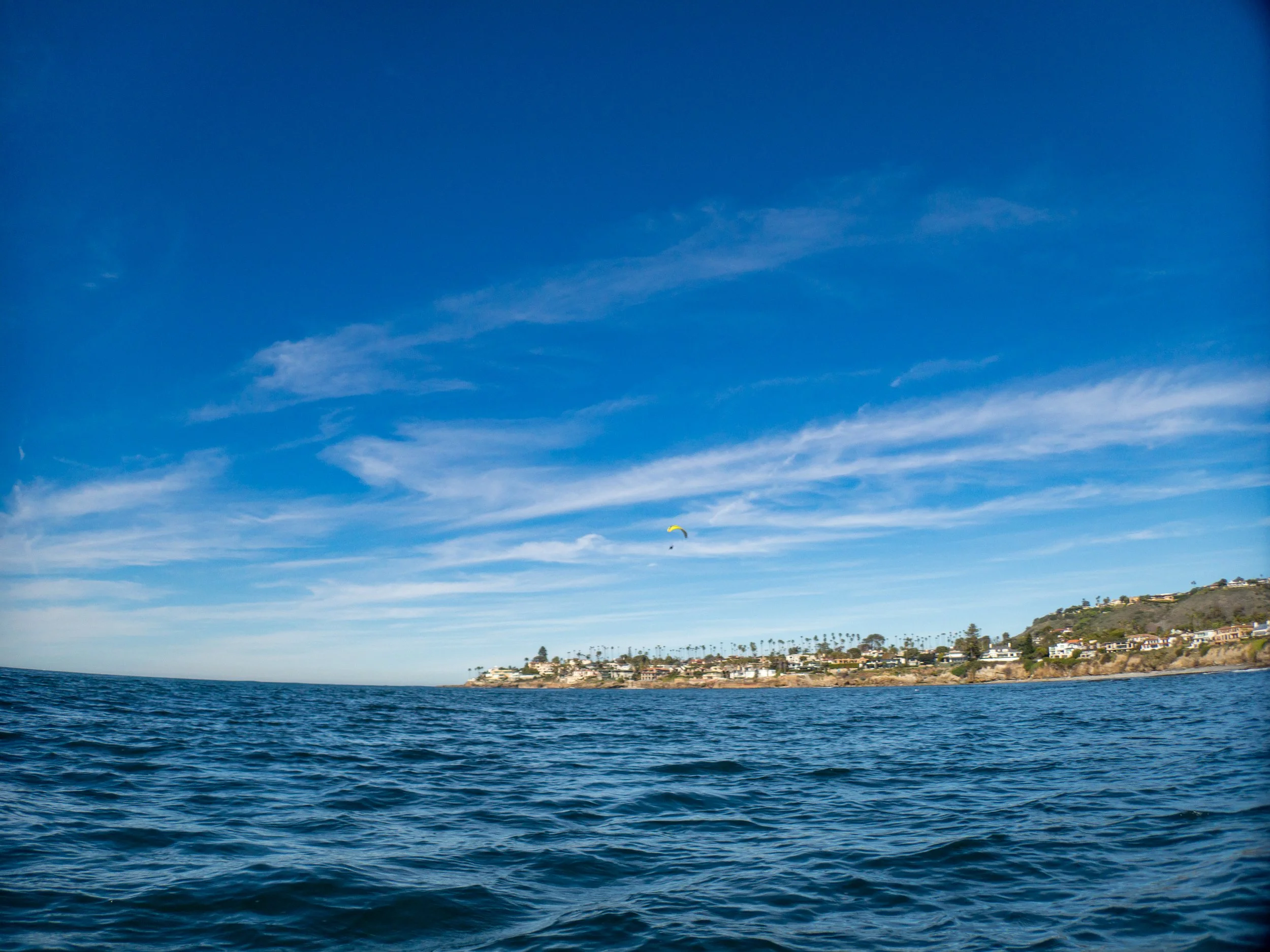 Ocean view with waves in the foreground, a coastal town with houses and palm trees on a hillside in the background, and a yellow parasail in the sky underneath wispy clouds.