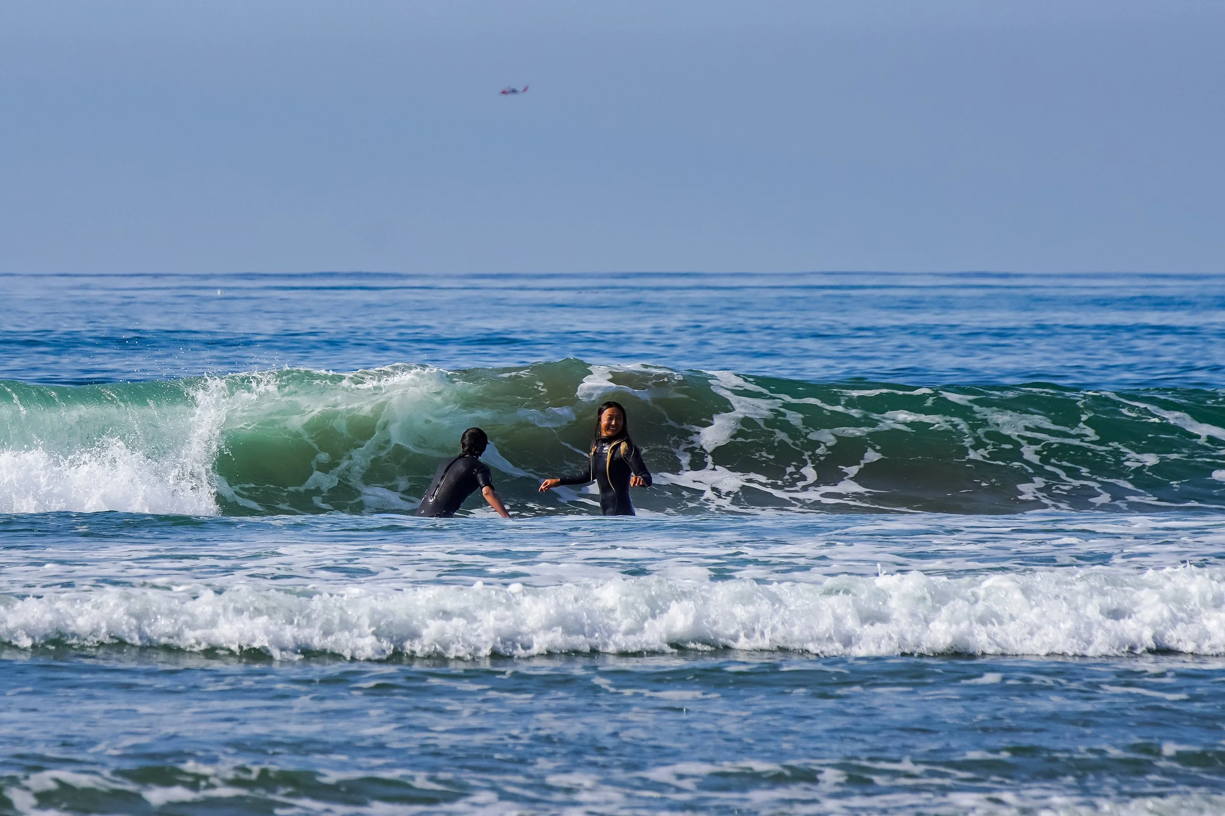 Two women in wetsuits playing in the ocean waves, with one woman smiling and holding hands of the other, as a large wave approaches.