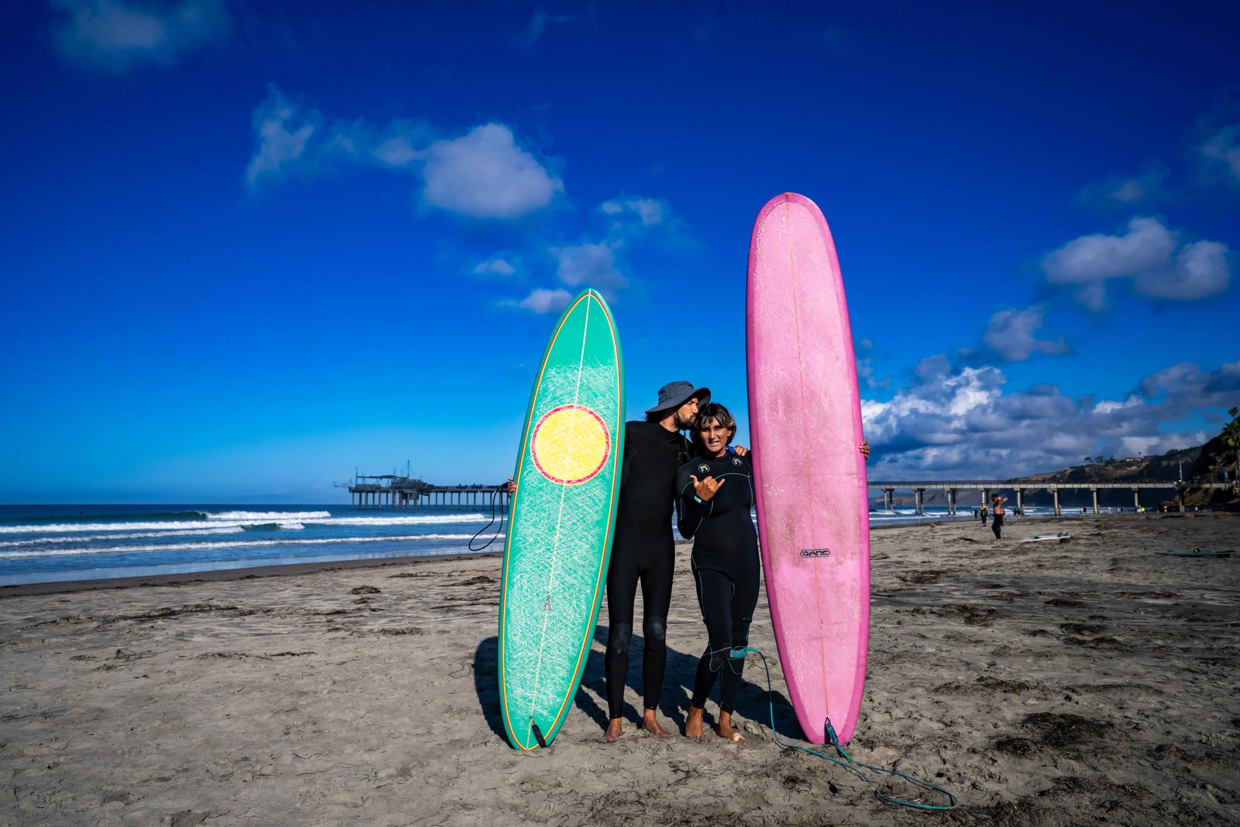Two people in wetsuits holding surfboards on a beach with blue sky and clouds, a pier in the background, and another person in the distance.