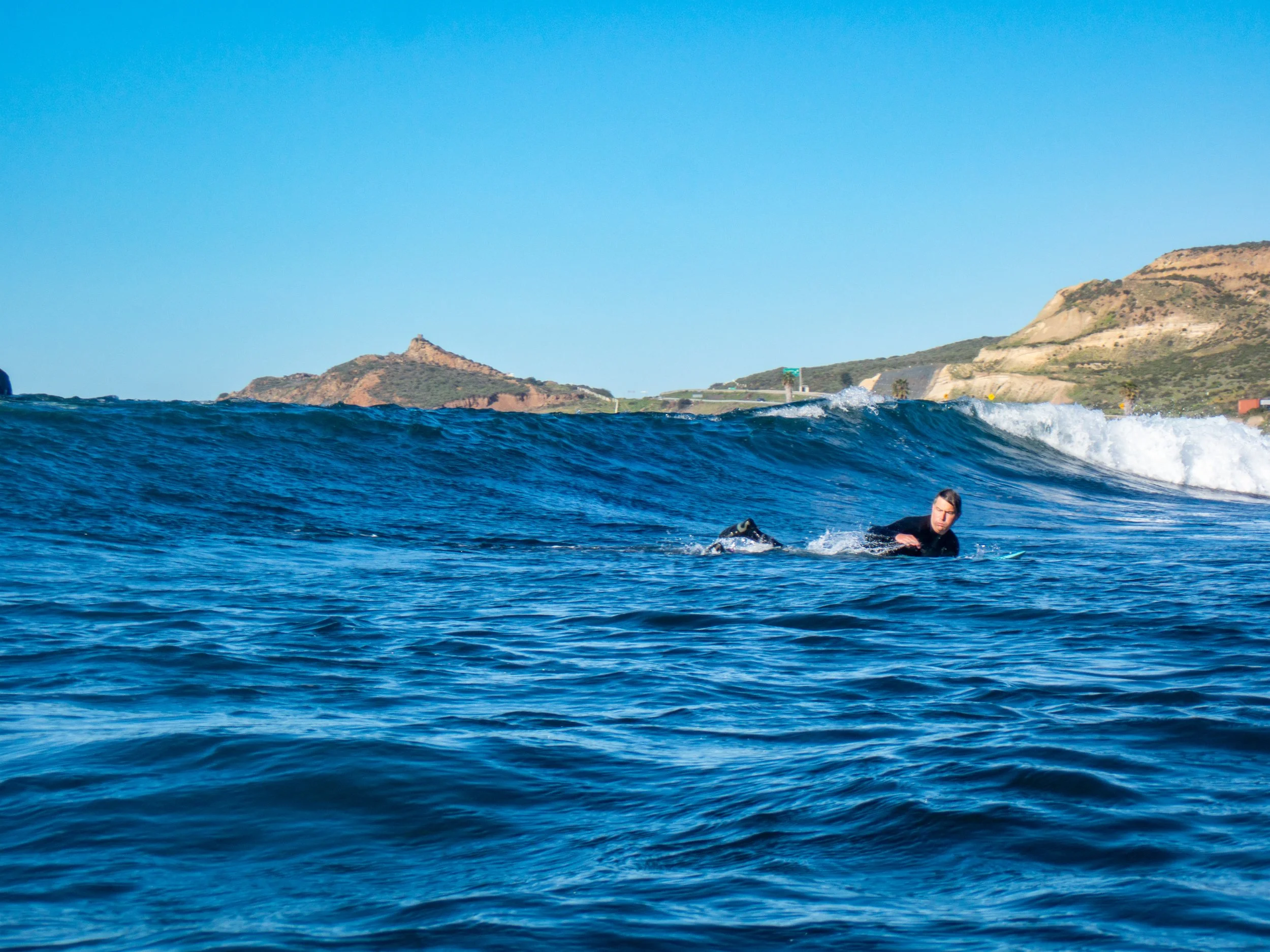 A person in a wetsuit lying on a surfboard in the ocean, with waves and distant hills under a clear blue sky.