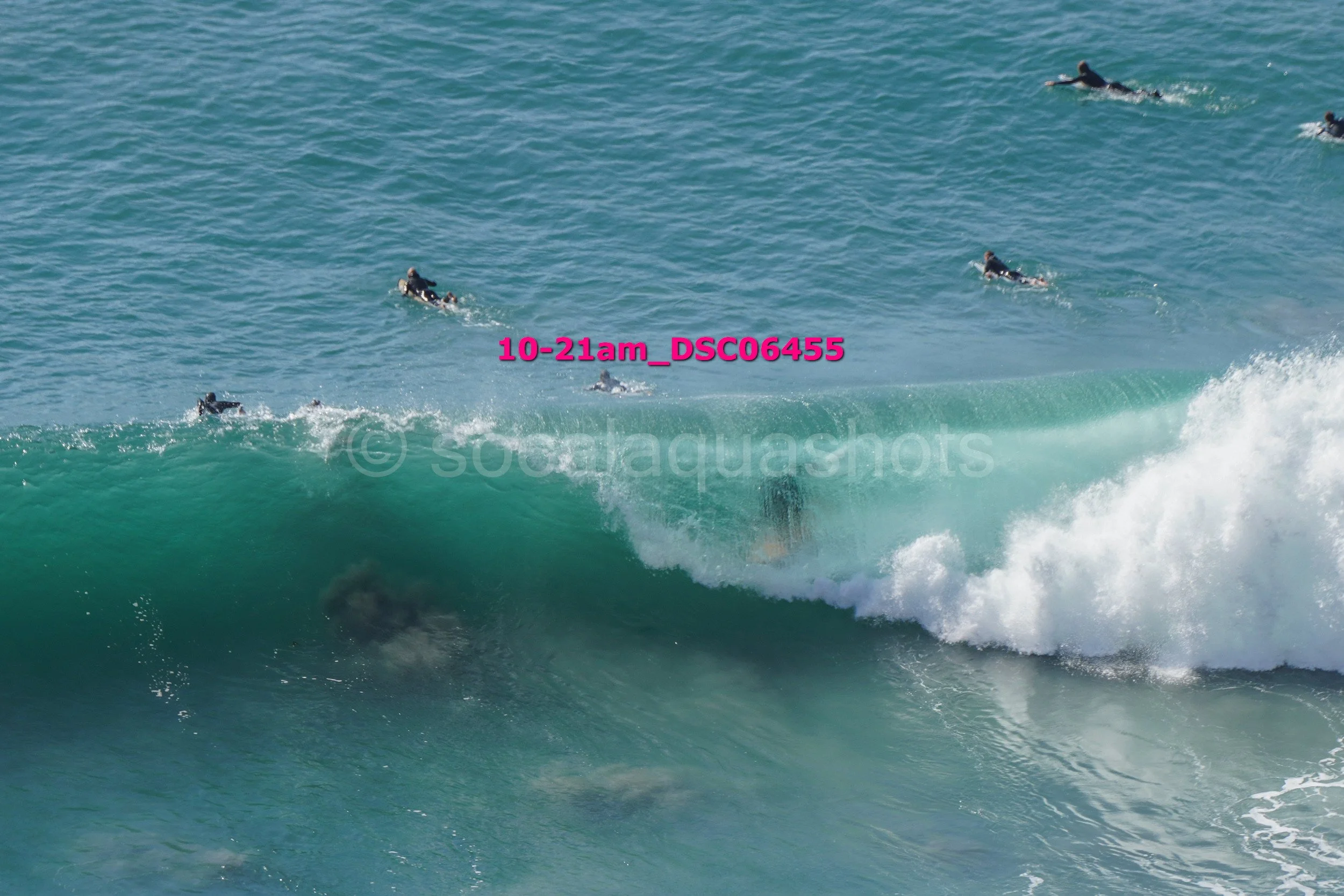 Surfers paddling out into the ocean on surfboards, with a large wave approaching. The water is clear and blue-green, with white foam at the wave's crest.