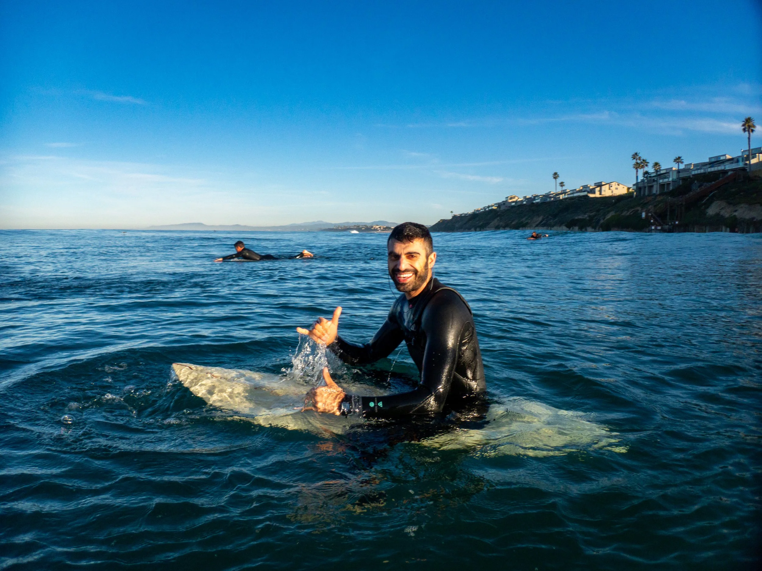 Smiling man in a wetsuit giving a thumbs up while sitting on a surfboard in the ocean, with two other surfers in the background and a coastline with houses and palm trees in the distance.
