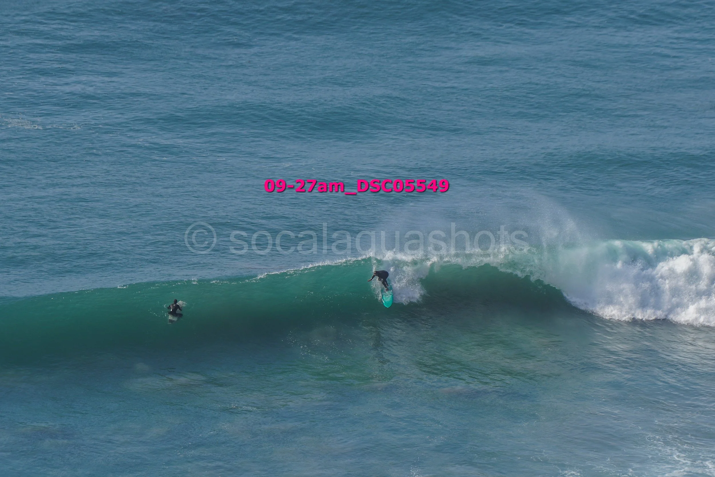 A person surfing on a turquoise wave with another person watching from the water