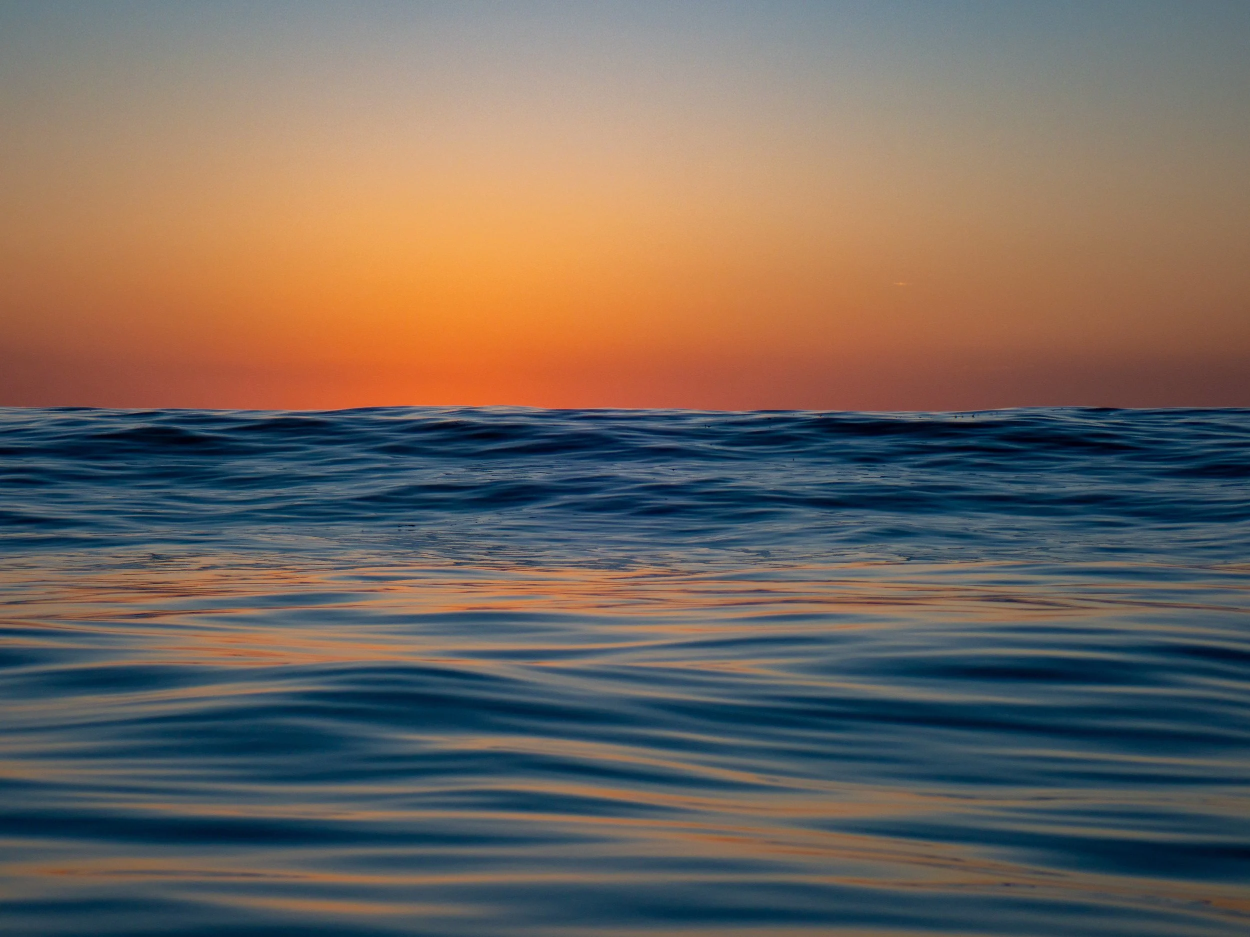 Calm ocean at sunset with horizon and vibrant orange sky reflected on water.
