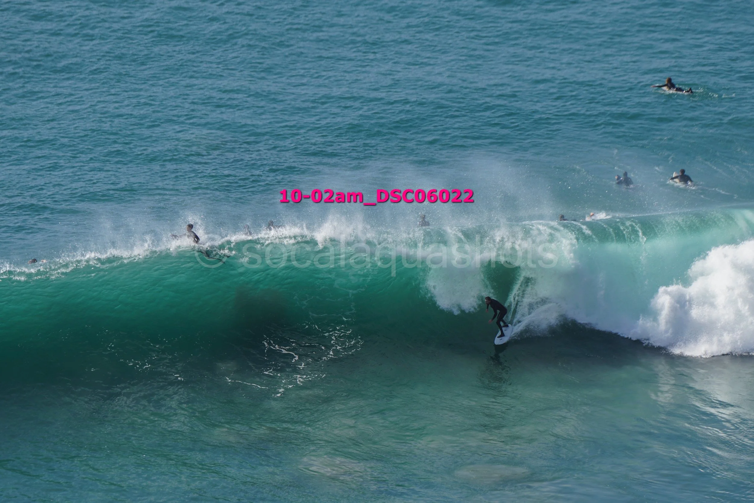 A surfer riding a large wave while other surfers wait in the water in the background.