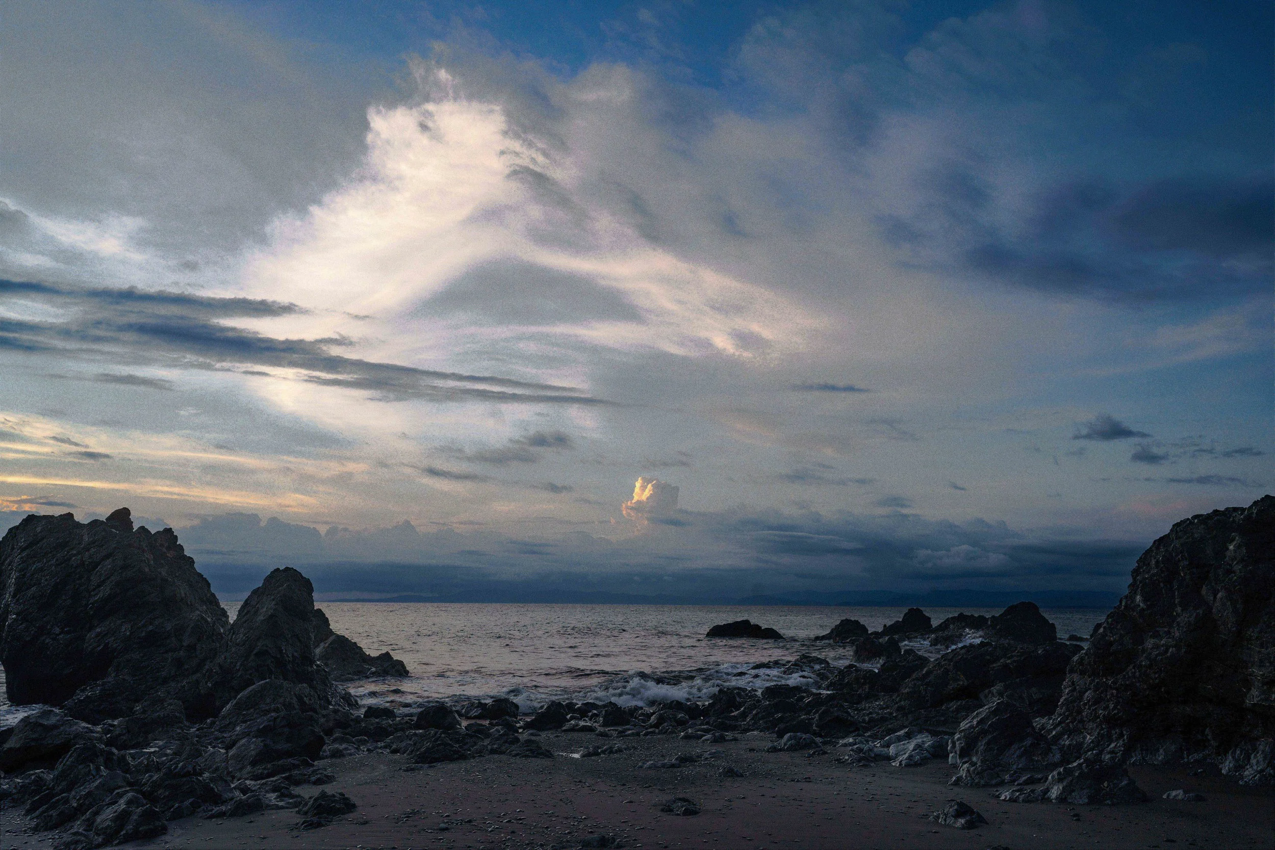 A rocky shoreline at sunset with dark clouds and a mostly cloudy sky over the ocean.