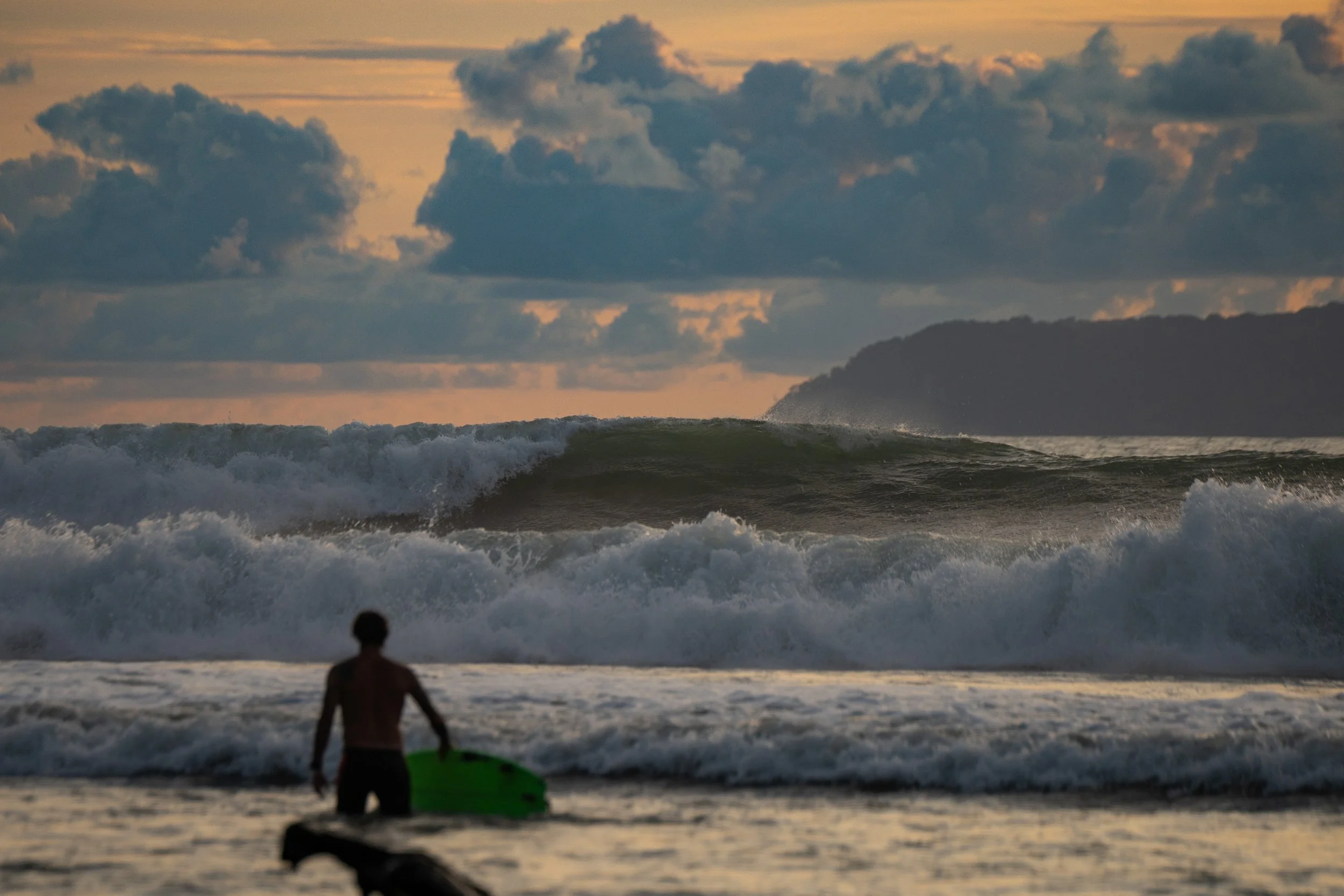 A person carrying a surfboard walks towards the ocean at sunset, with large waves and a cloudy sky in the background.