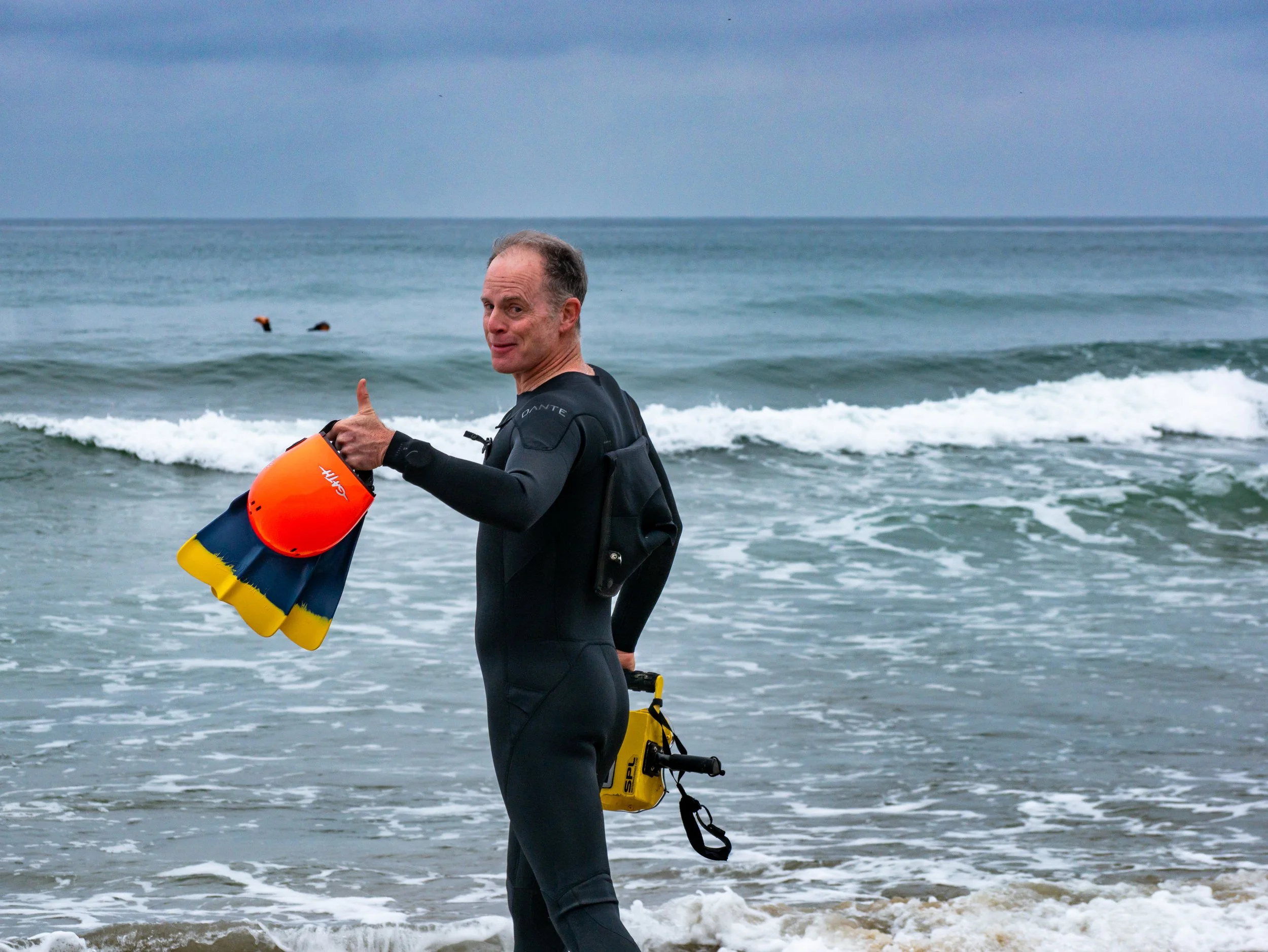Man in wetsuit holding a colorful helmet and giving a thumbs-up at the beach with ocean waves in the background.