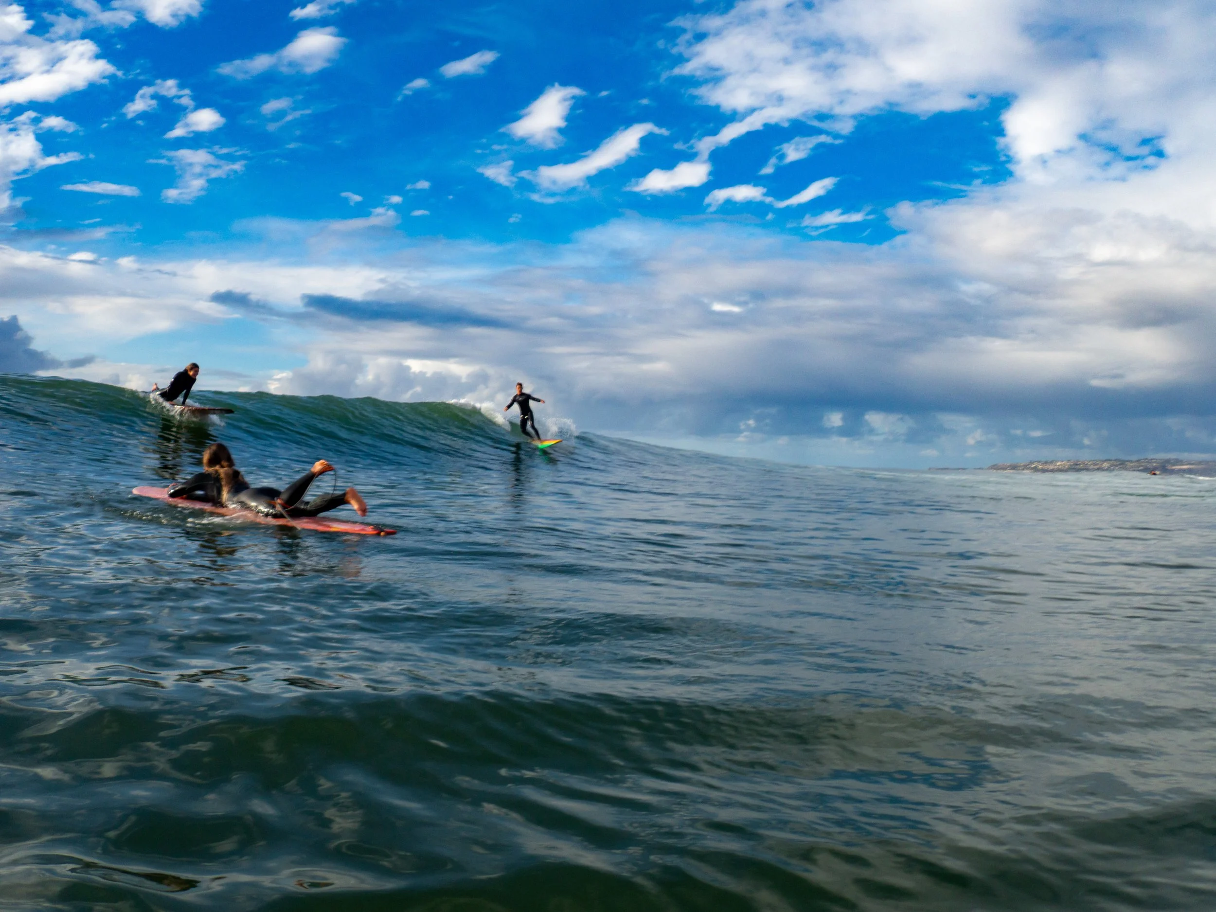 Three surfers riding or lying on surfboards on the ocean, with blue sky and clouds in the background.