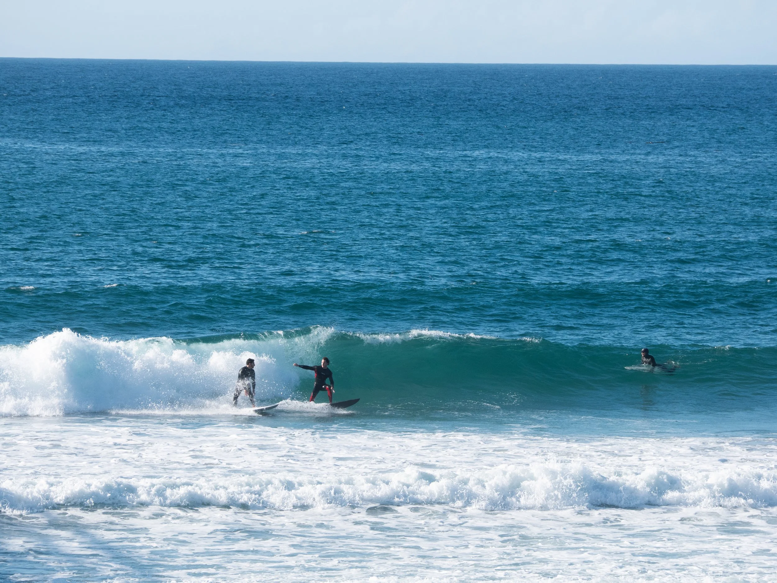 Three people surfing on a blue ocean wave, two standing on surfboards and one paddling in the water.