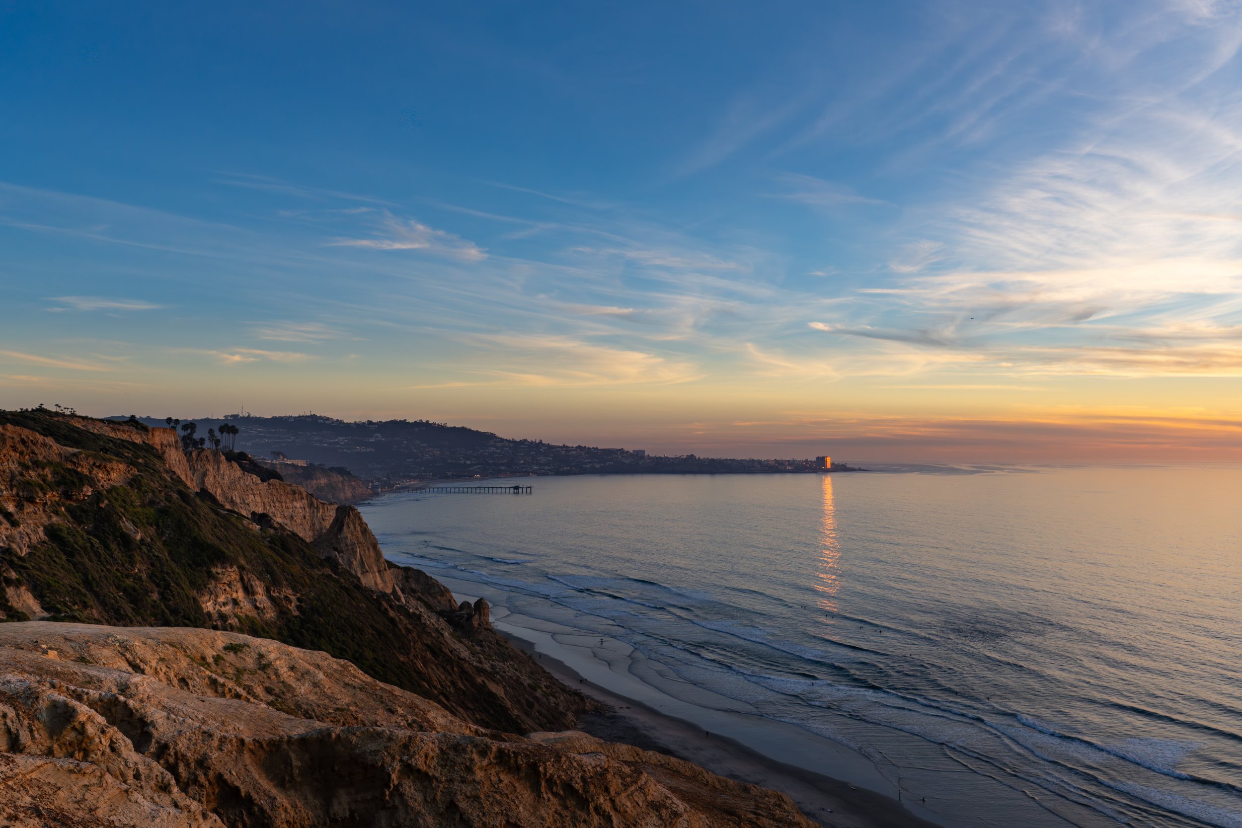 Coastal cliffs overlooking an ocean at sunset with a pier extending into the water and a distant city skyline.