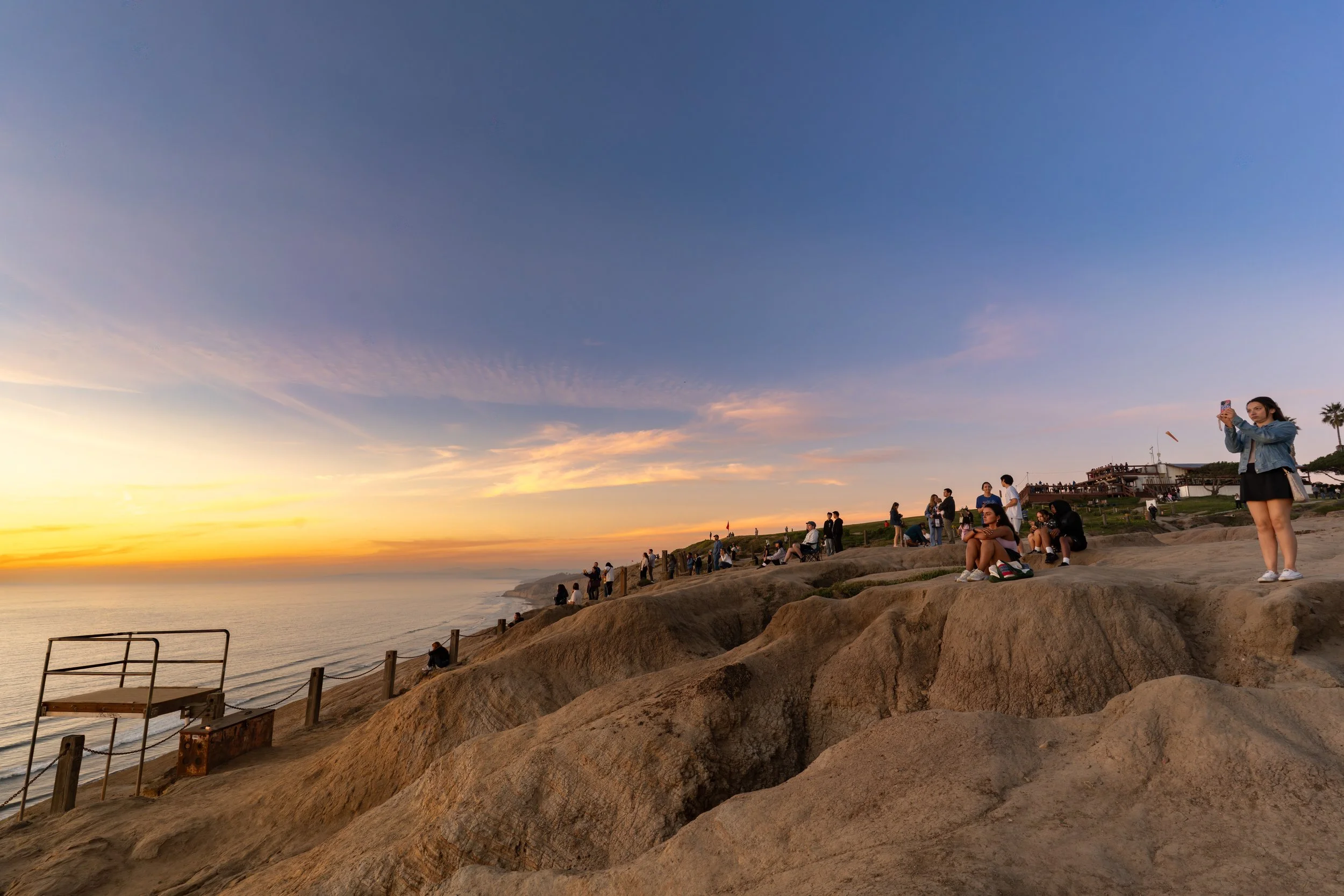 People sitting and standing on rocky cliffs along the coast during sunset, some taking photos and enjoying the view.