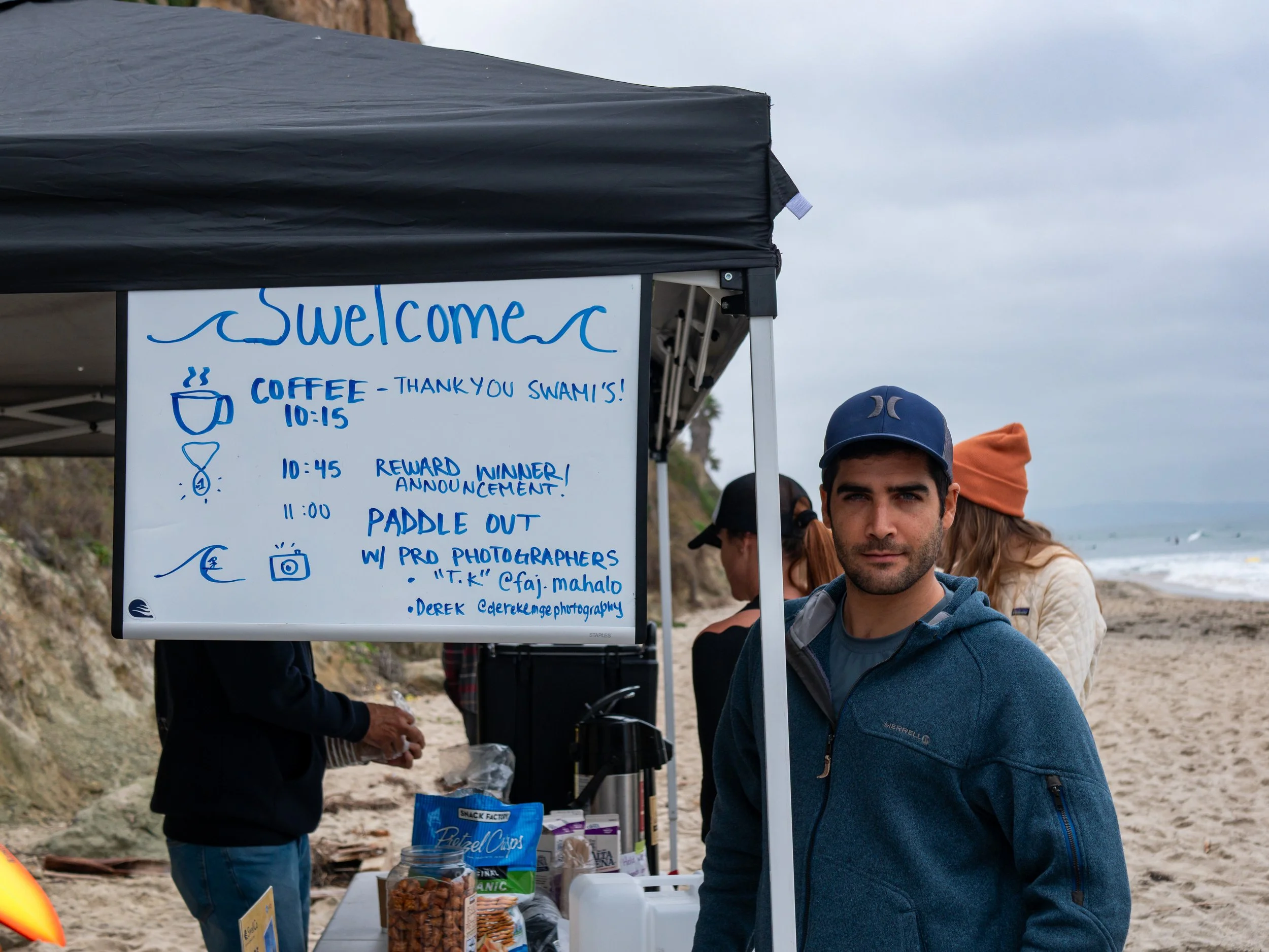A man in a blue jacket and cap standing at a beach-side refreshment stand near a whiteboard sign with schedule details for an event, with people and the ocean in the background.
