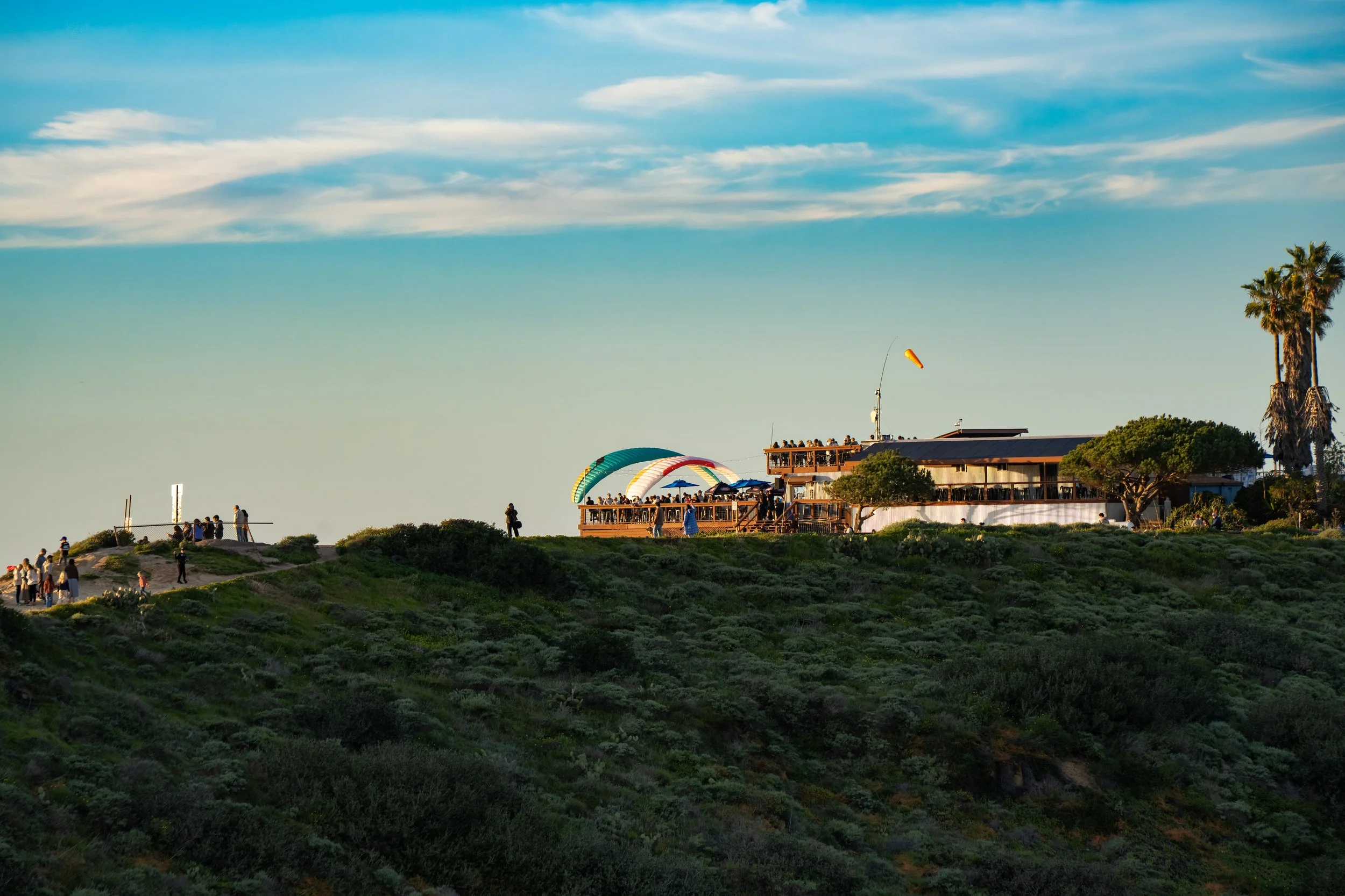 People gathered on a wooden deck overlooking a green hillside, with a building, palm trees, and a wind sock in the background, under a blue sky with some clouds.