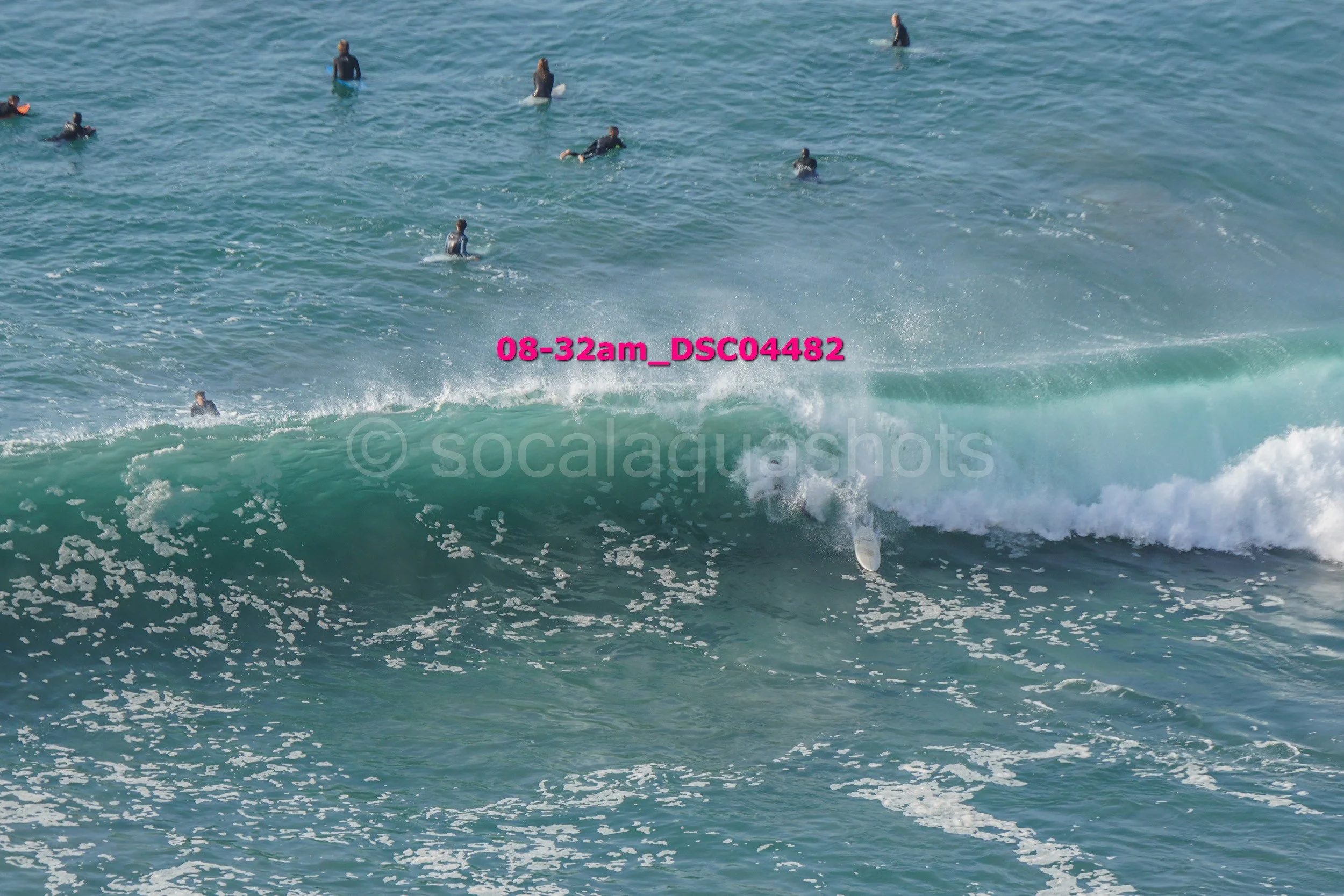 Surfer falling off a wave in the ocean while multiple people are in the water, some standing and some swimming.
