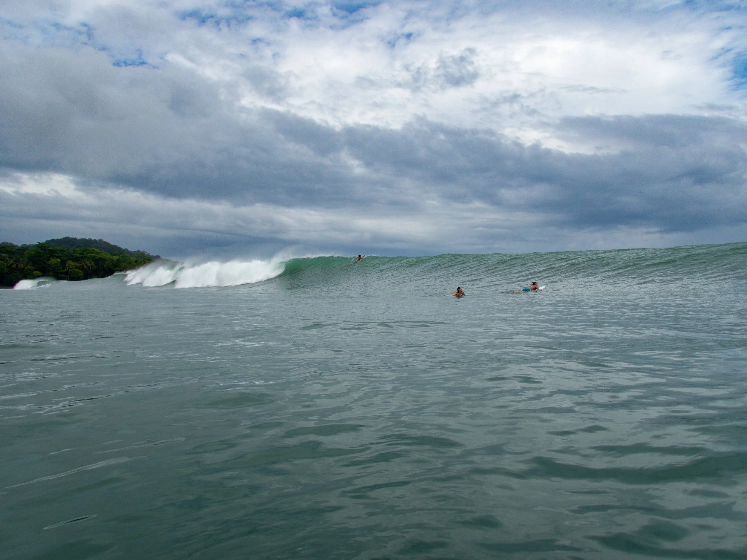 Large ocean wave with surfers paddling, cloudy sky, coastal view in background.