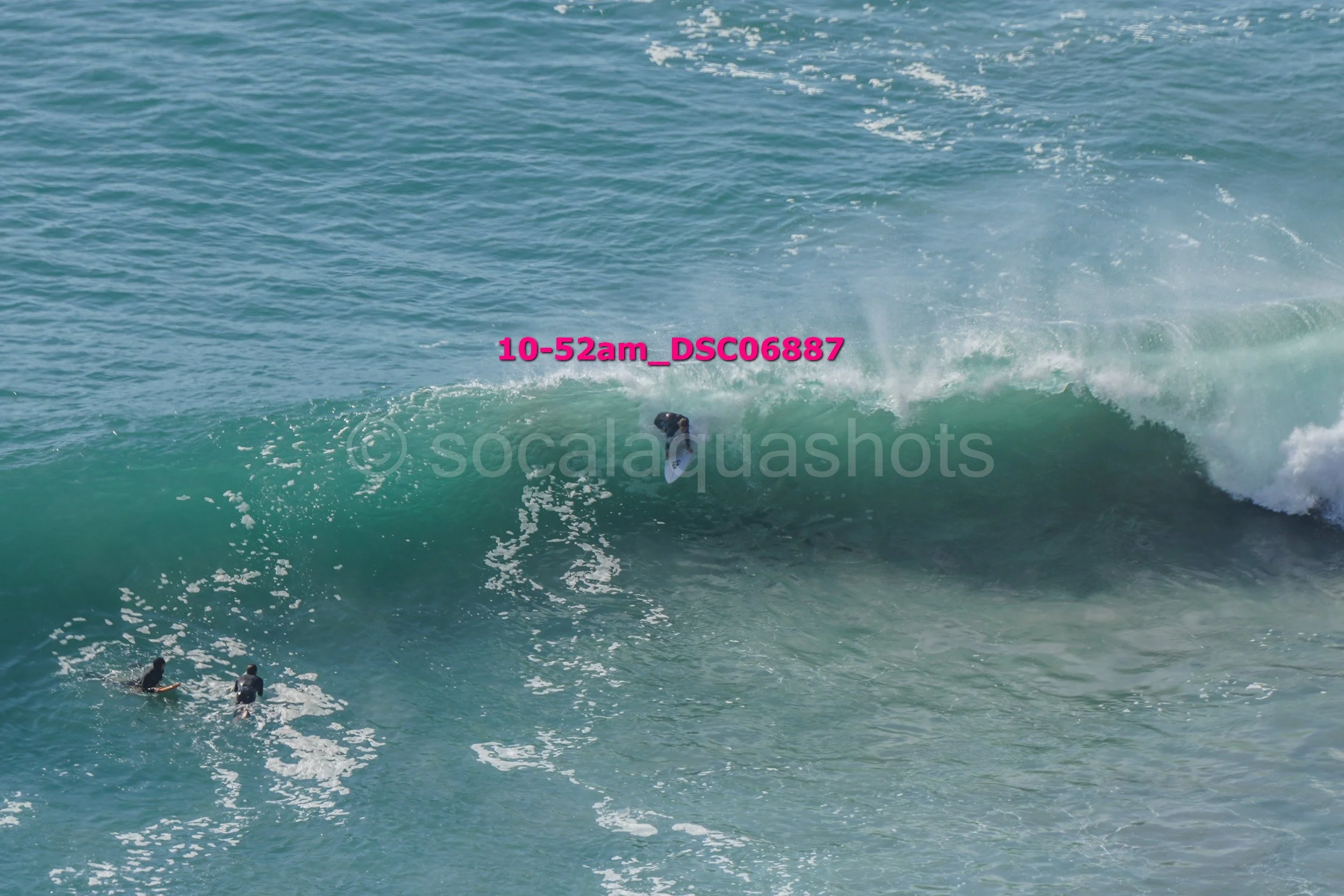 A person surfing on a large ocean wave with two surfers paddling nearby.
