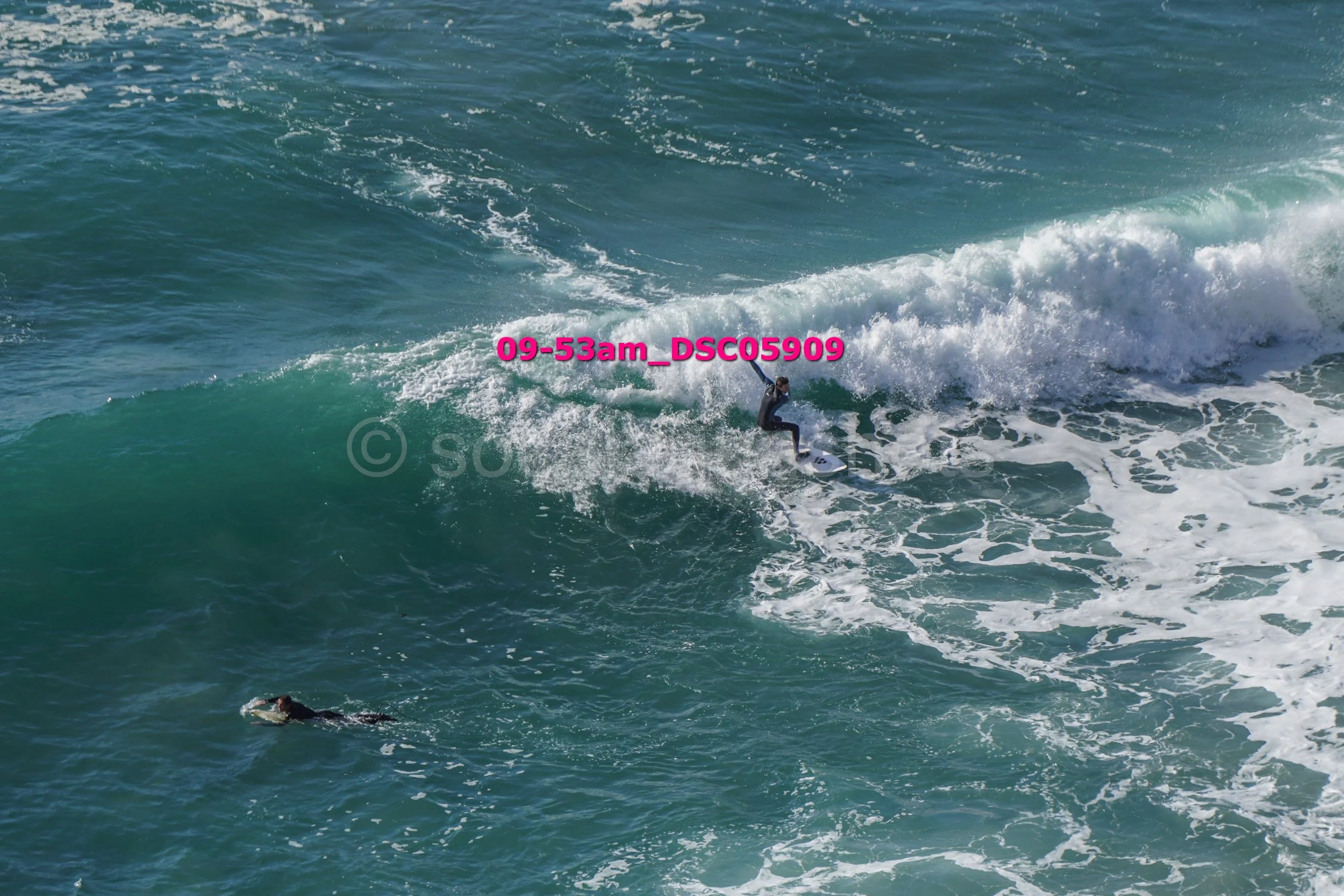 A person surfing on a large wave in the ocean with another surfer in the water nearby.
