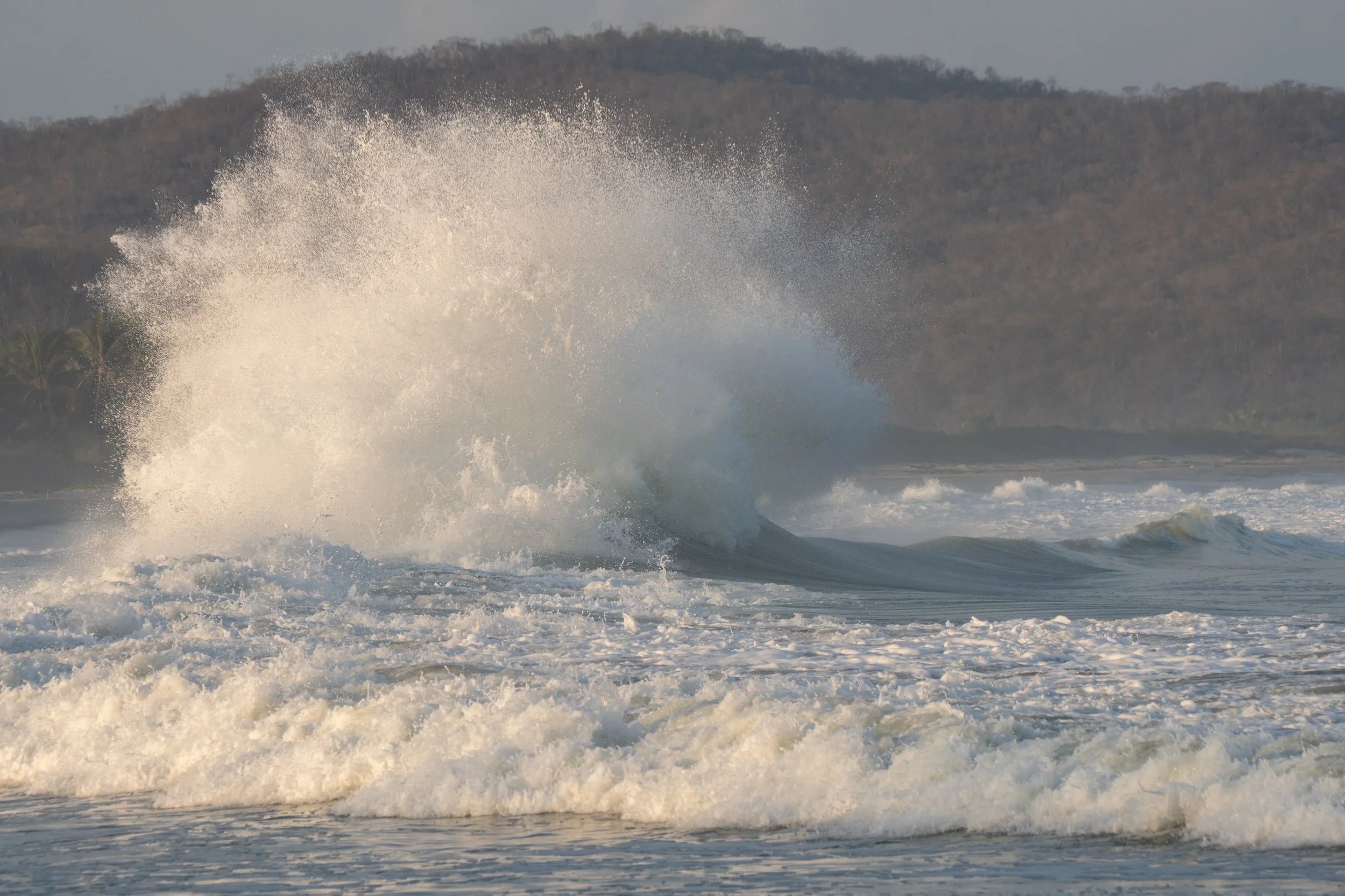 Waves crashing in the ocean with a hill in the background, overcast sky.