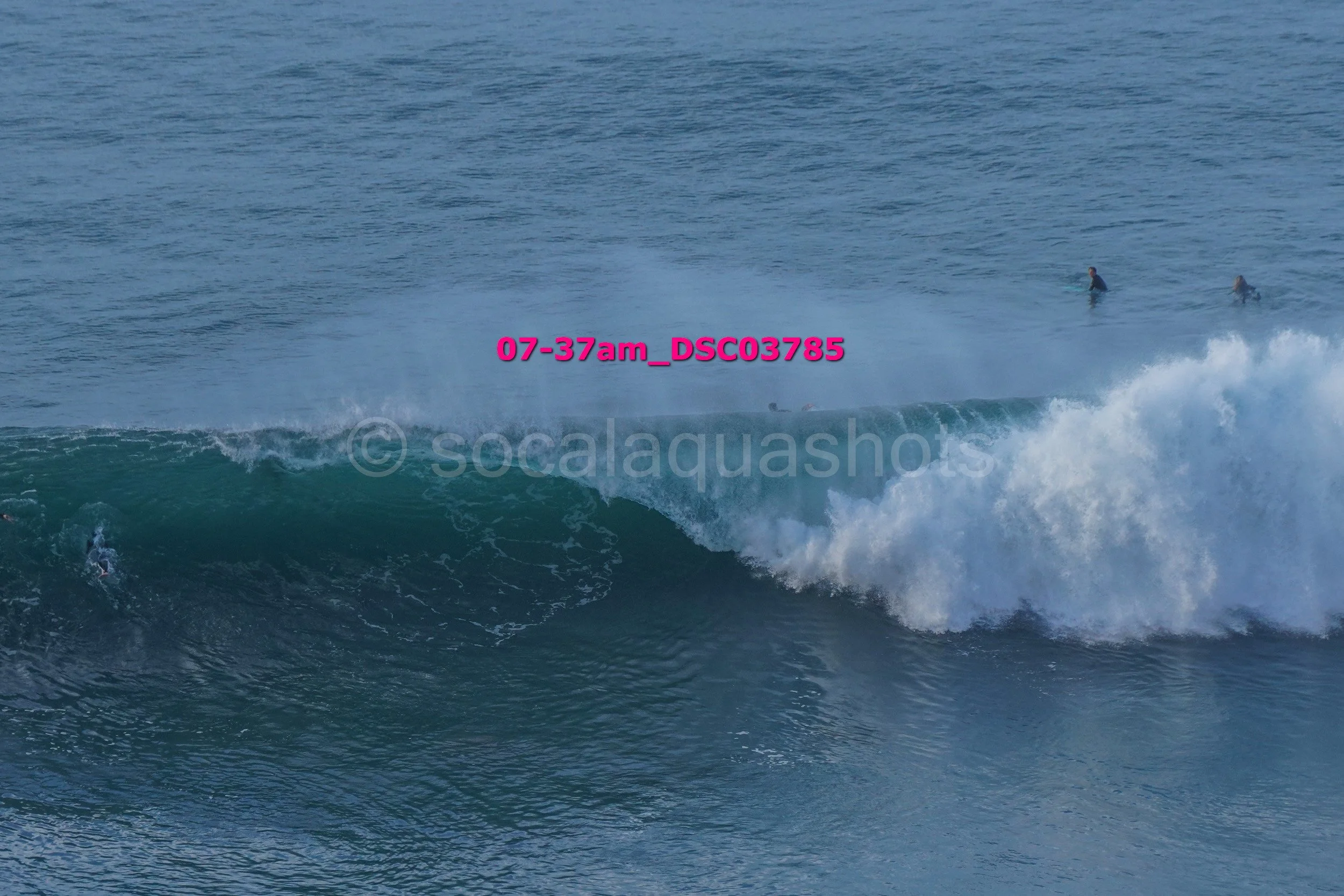 View of ocean waves with surfers in the water, some riding waves and others waiting, in a beach scene.