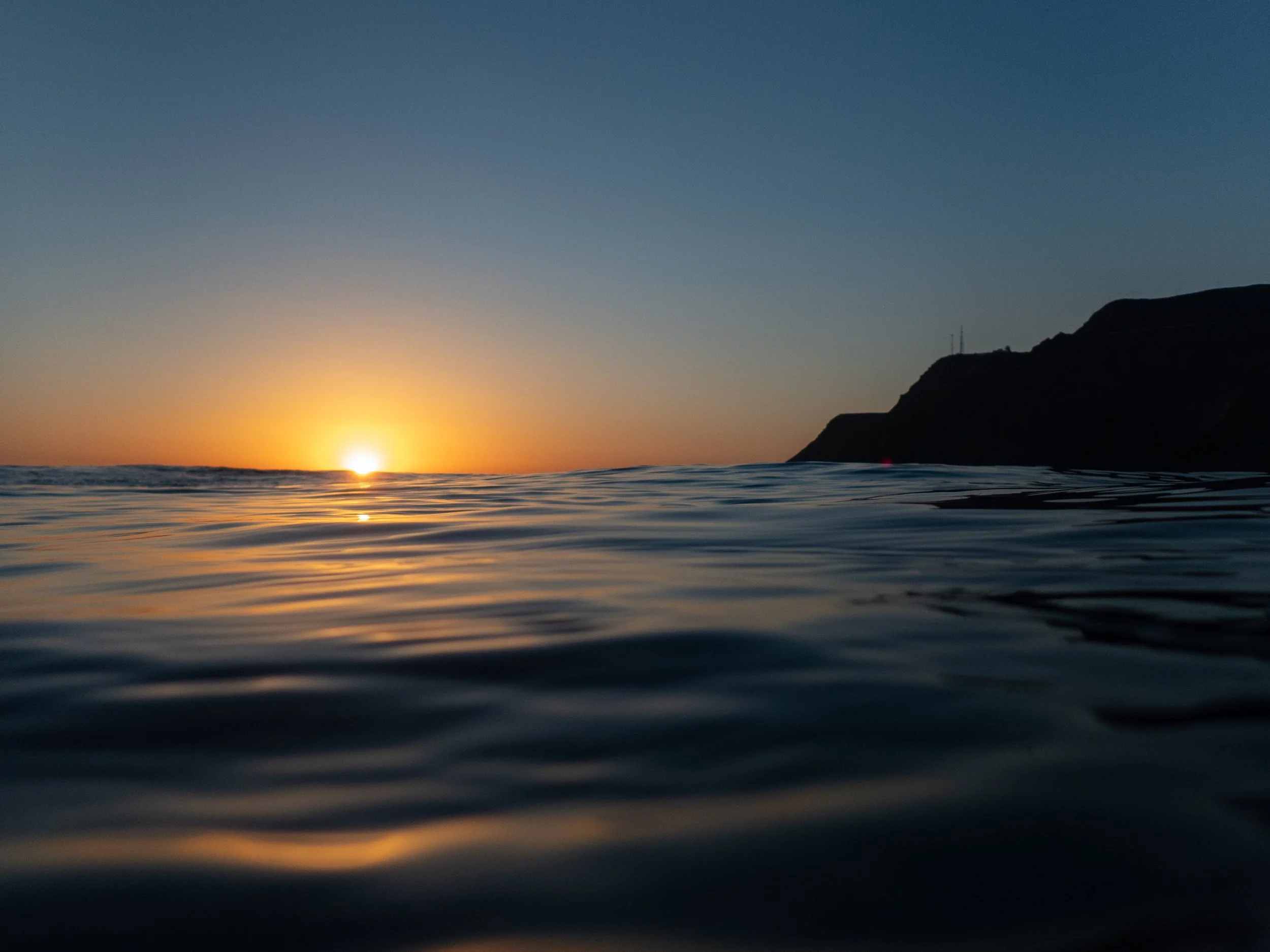 Photo of the sun setting over the ocean with gentle waves and a silhouetted coastline on the right.