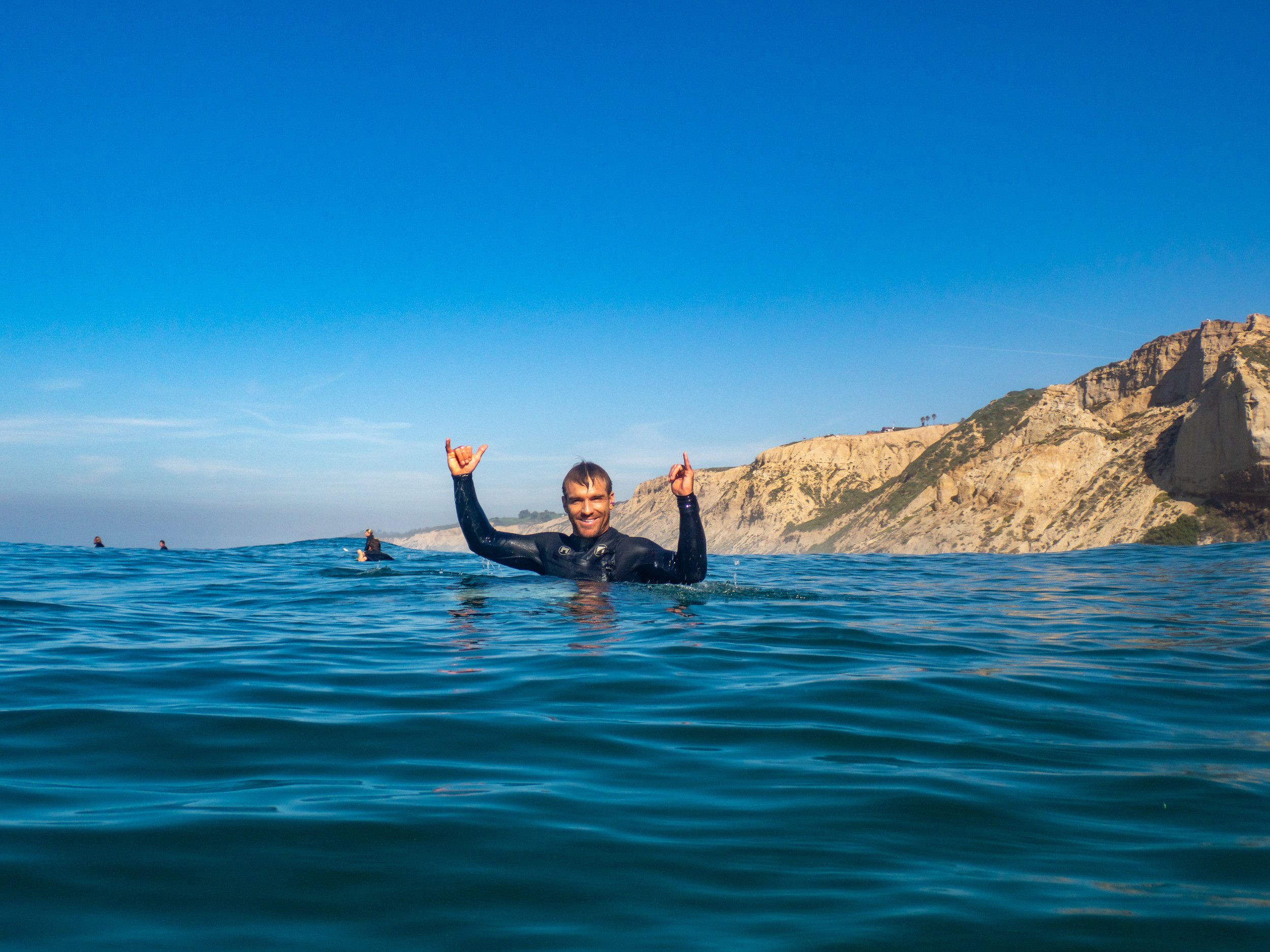 Person in wetsuit swimming in the ocean near cliffs, smiling and making a shaka sign with both hands, under a clear blue sky.