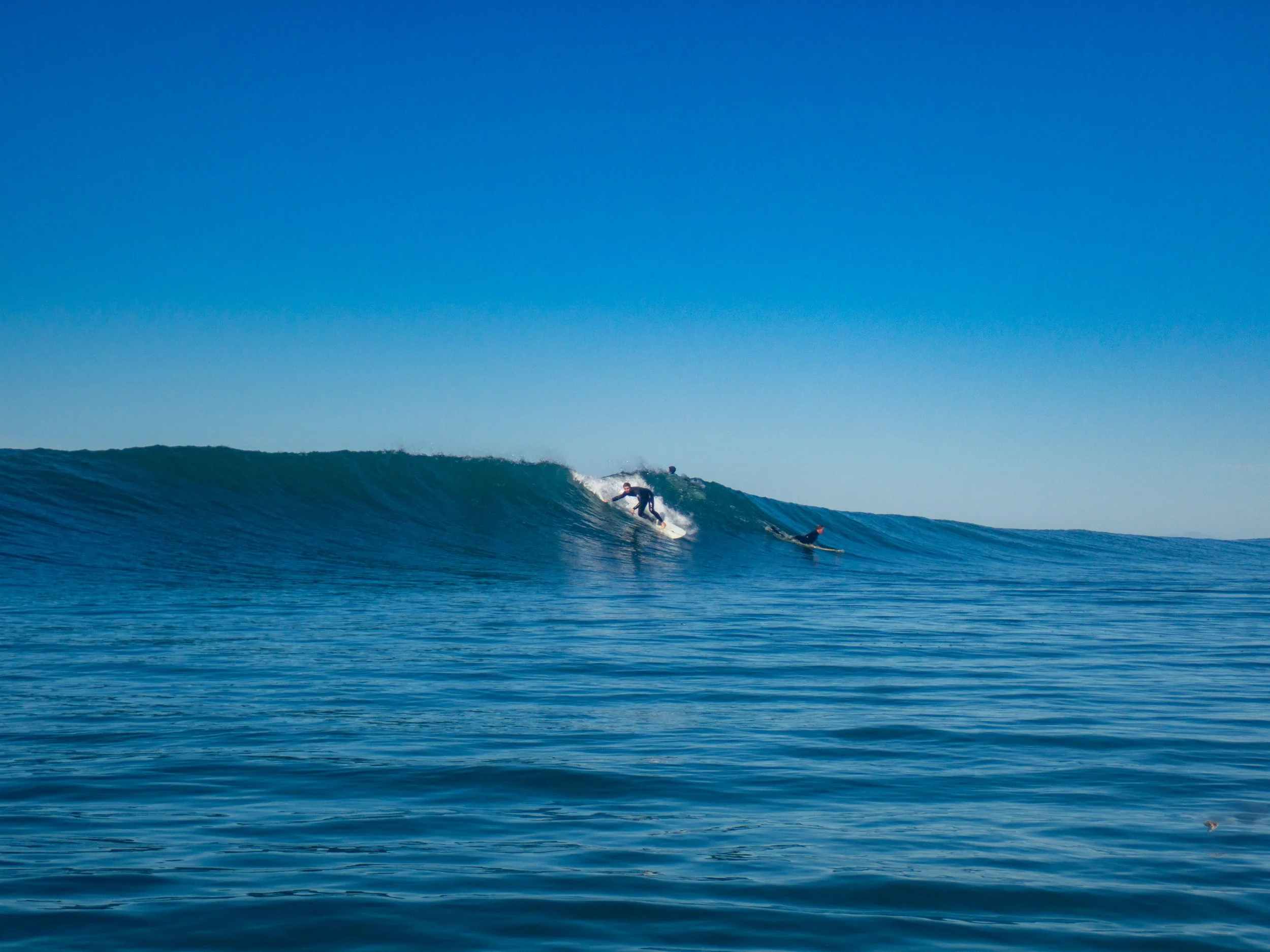 A person surfing on a large wave with two other surfers in the water under a clear blue sky.