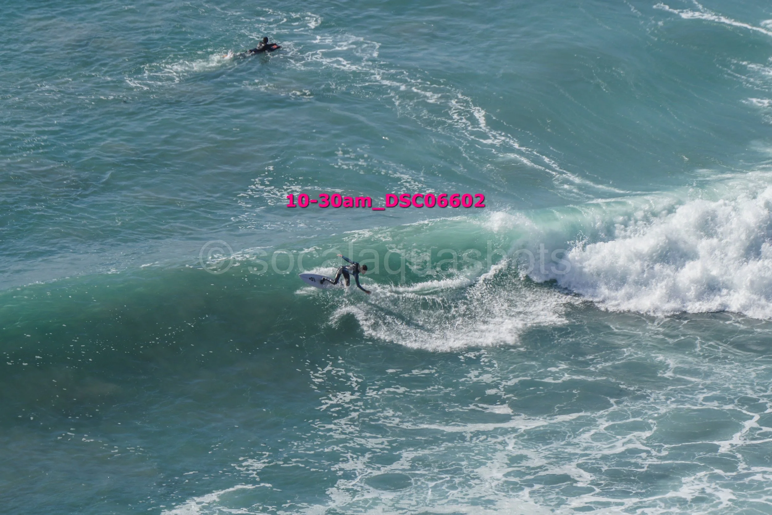 A person surfing on a wave in the ocean, with another person swimming or surfing further out.