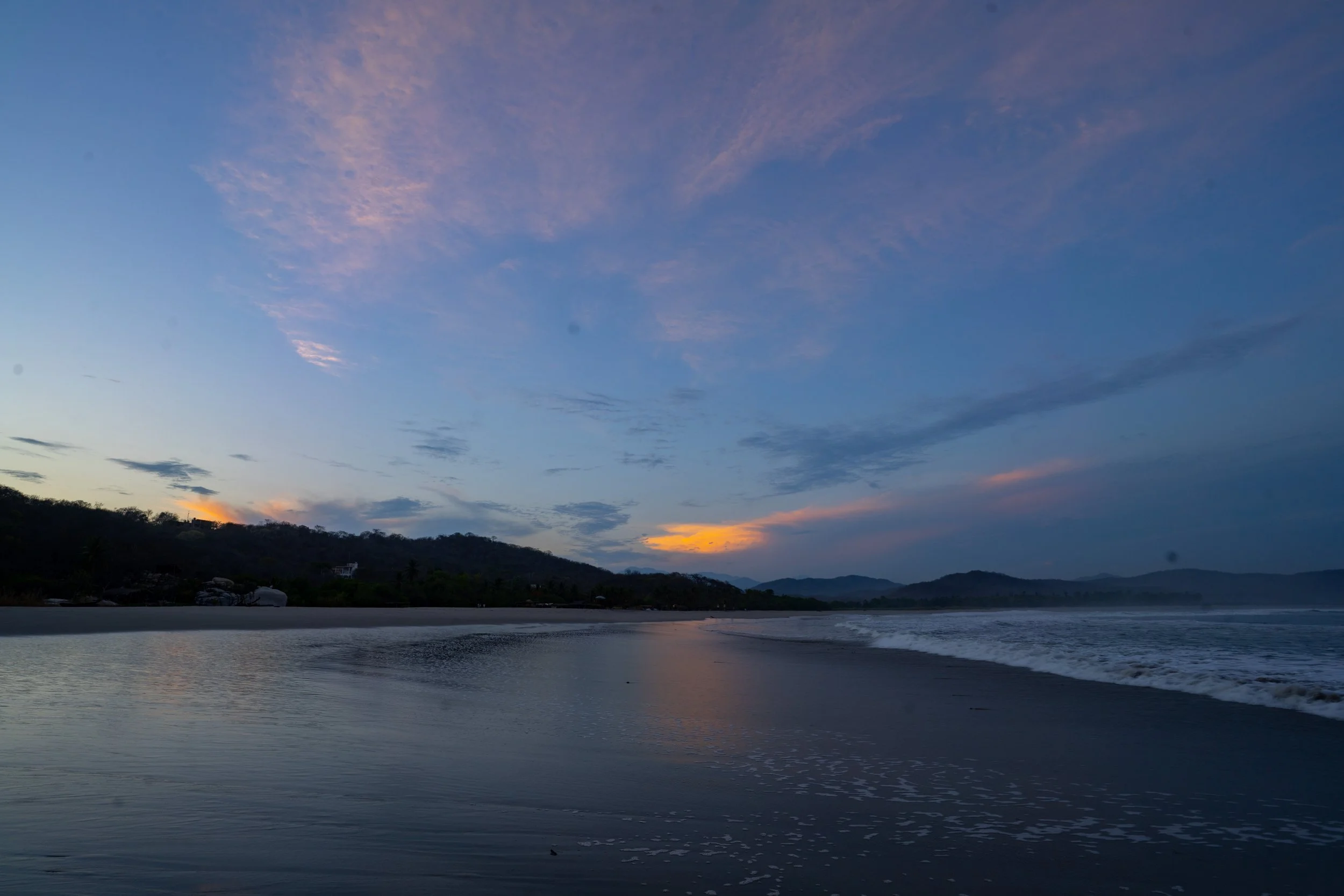 Sunset over a beach with calm water, distant mountains, and a partly cloudy sky with pink and orange hues.