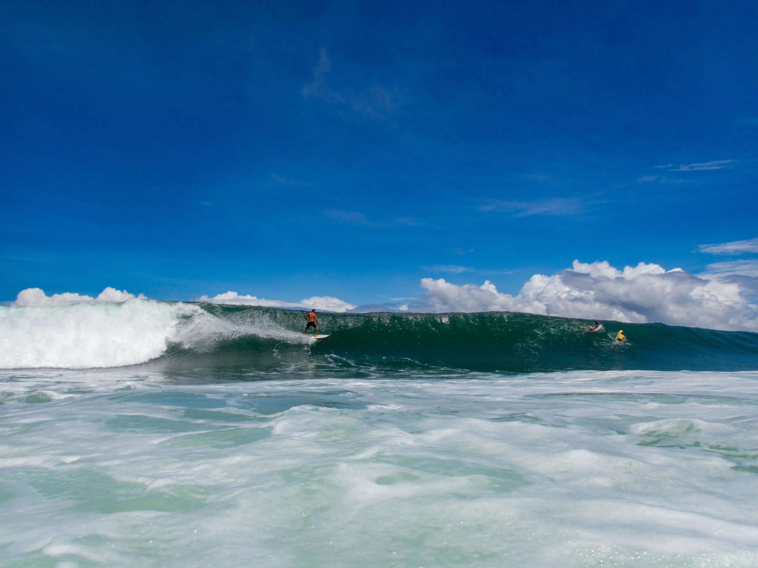 Surfers riding a wave in the ocean on a sunny day with blue sky and clouds.