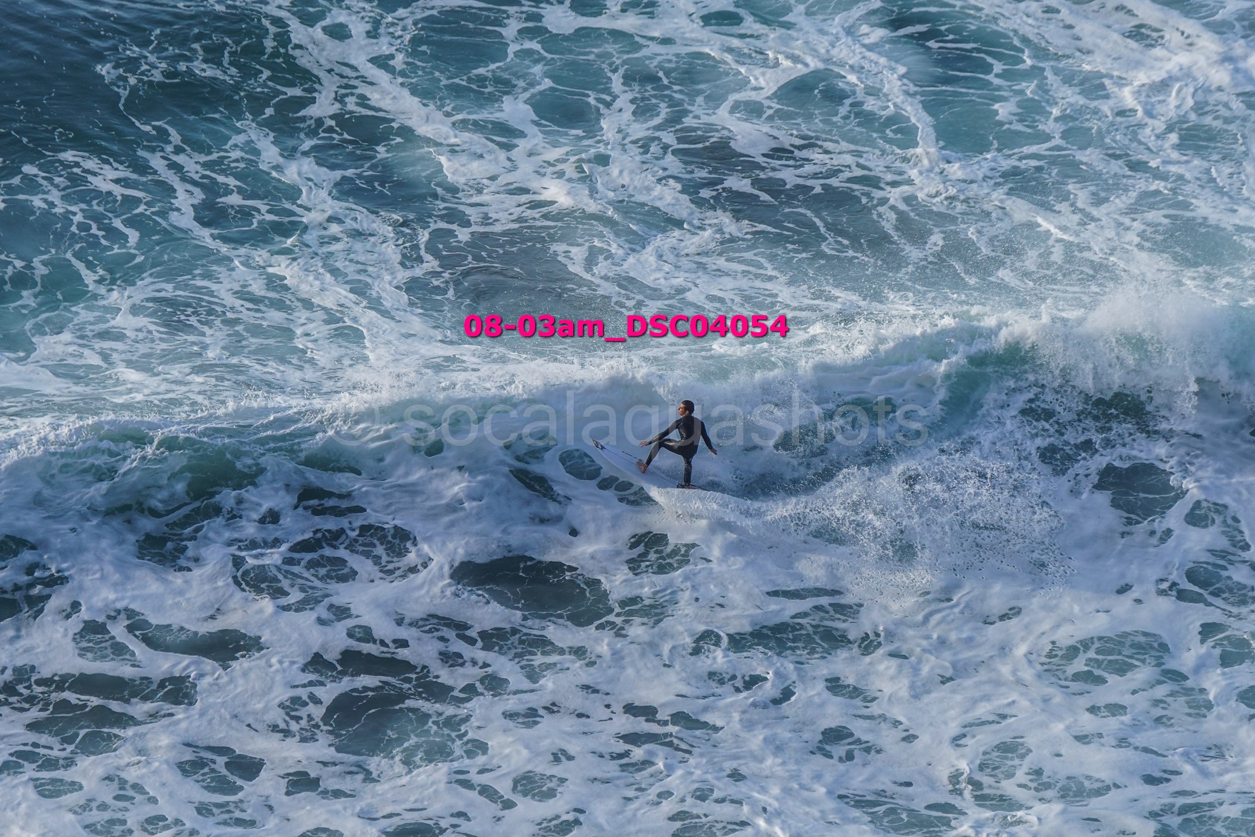 A person surfing on a wave in the ocean with white foam and blue water, seen from above.