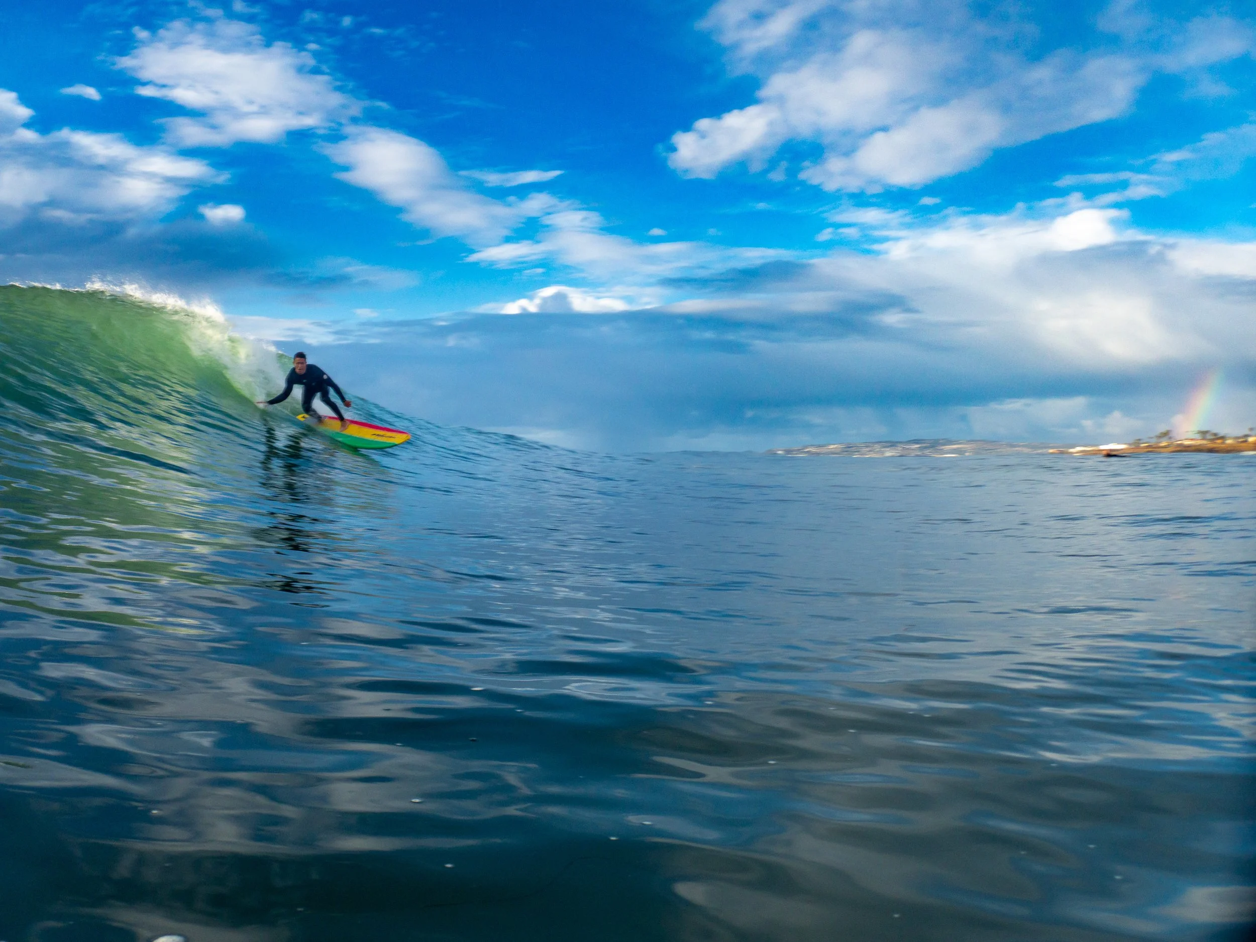 A person surfing on a green and yellow surfboard in the ocean with a blue sky, scattered clouds, and a faint rainbow in the background.