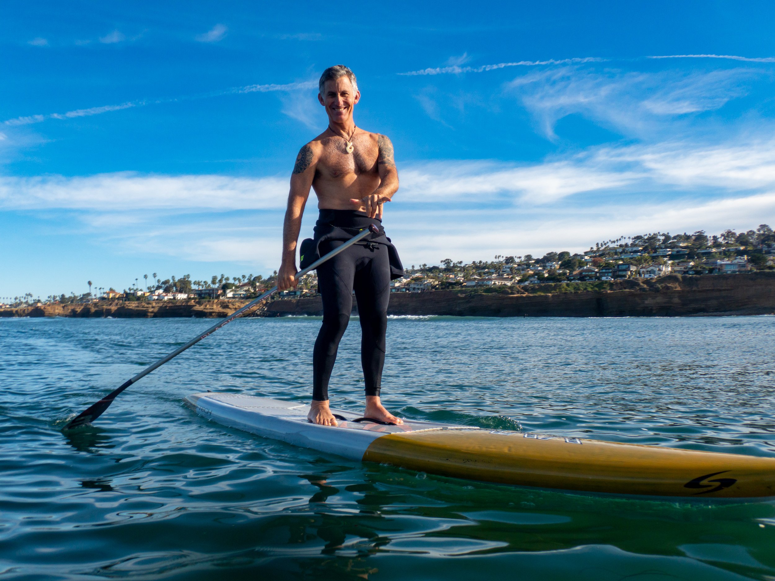 A shirtless man with tattoos on his arms standing on a paddleboard in the water, holding a paddle, with a coastline and houses in the background under a blue sky.