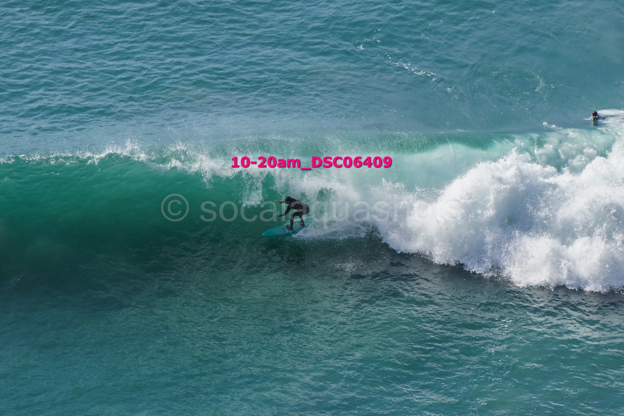 A person surfing on a large wave in the ocean, wearing a black wetsuit, with another surfer visible in the background.