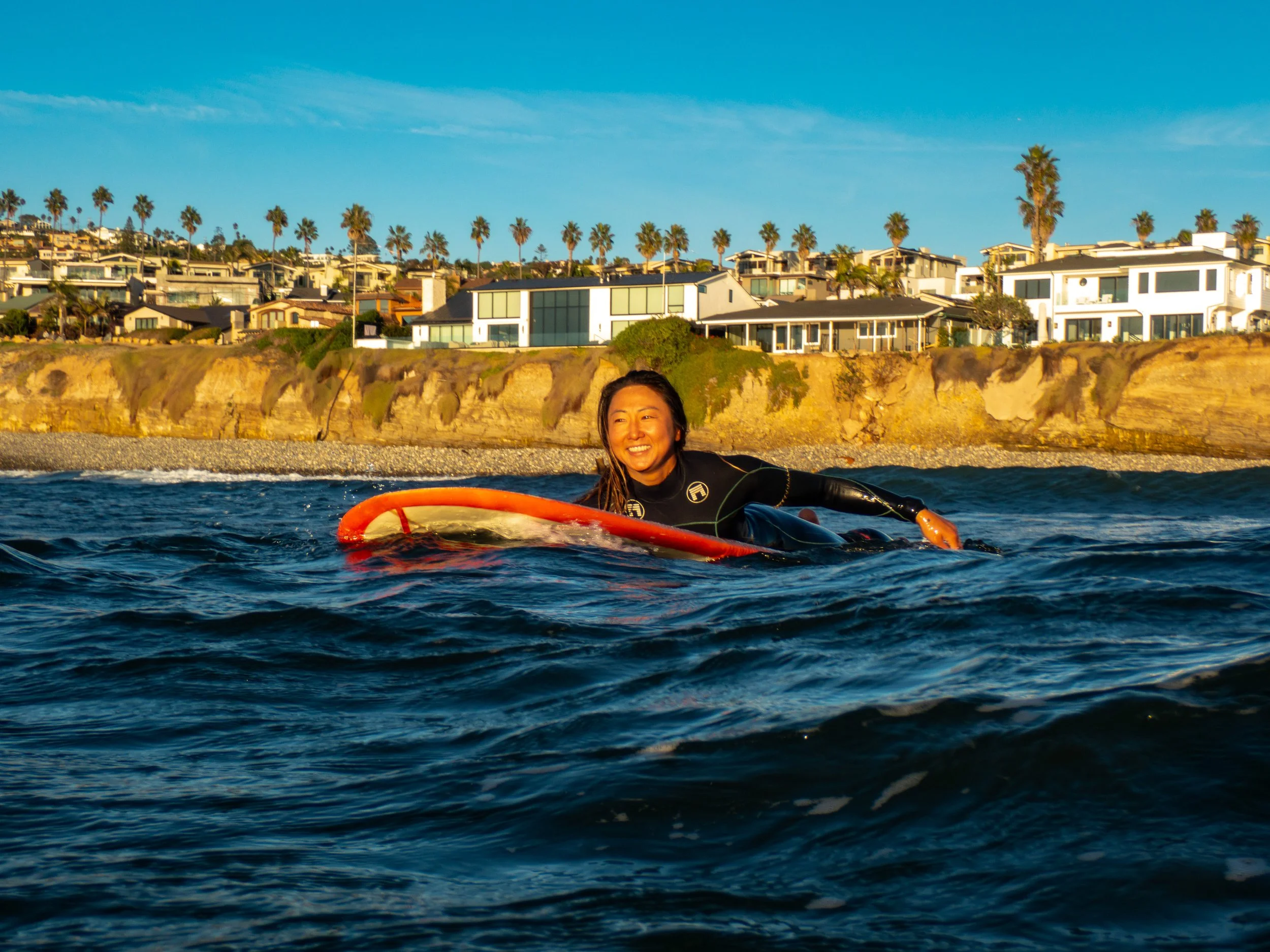 A woman surfing on a surfboard in the ocean with a coastal residential area and palm trees in the background during sunset.